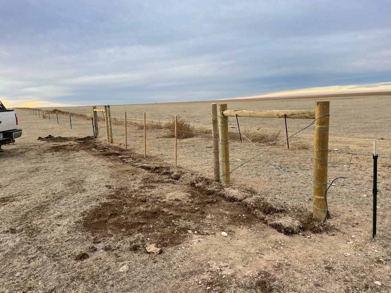 A newly installed barbed wire fence supported by wooden posts running across a flat, open, dry field under a cloudy sky, with part of a white truck visible on the left.