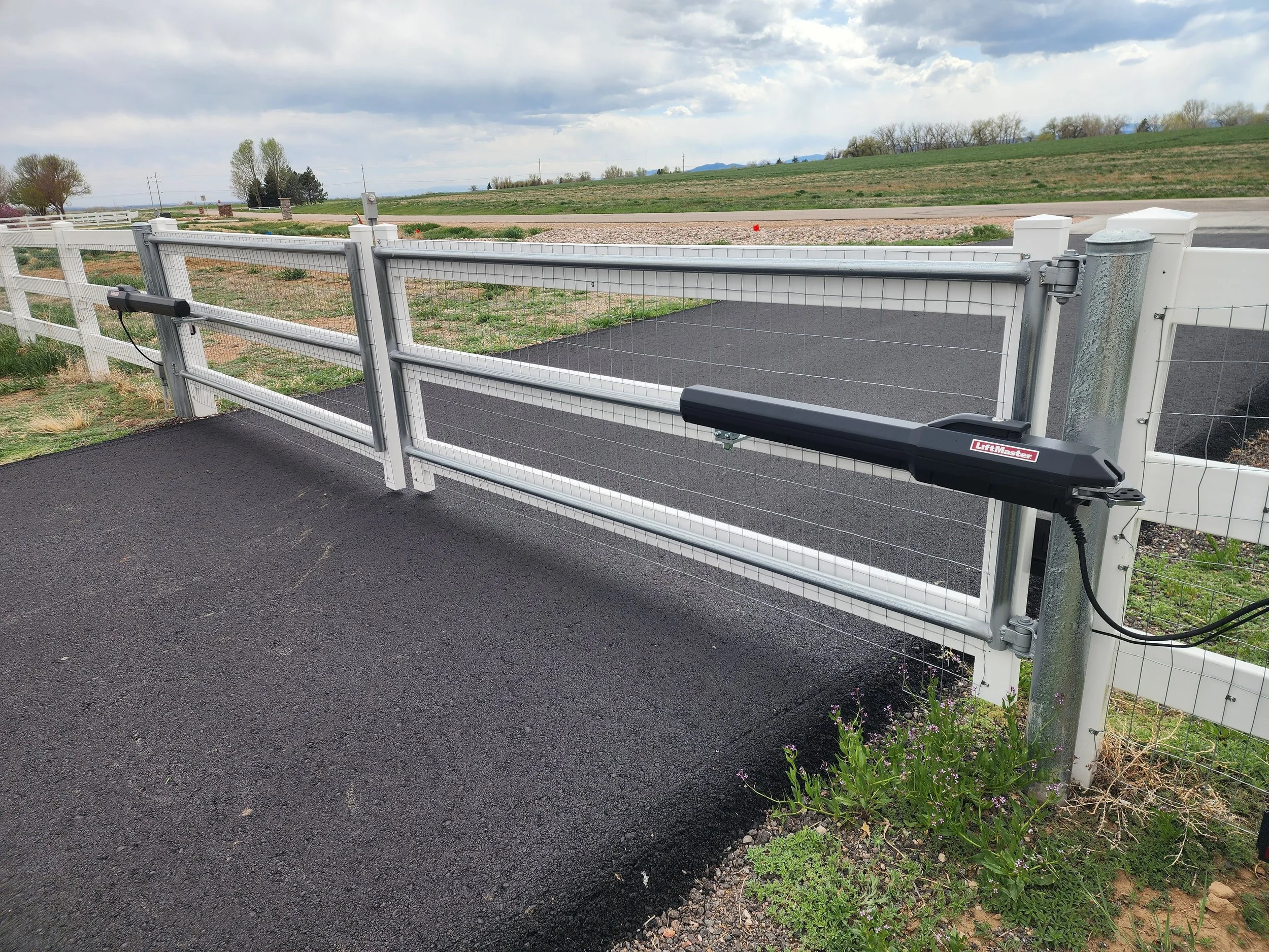 Automatic sliding gate with a driveway and a background of farmland and trees under a cloudy sky.