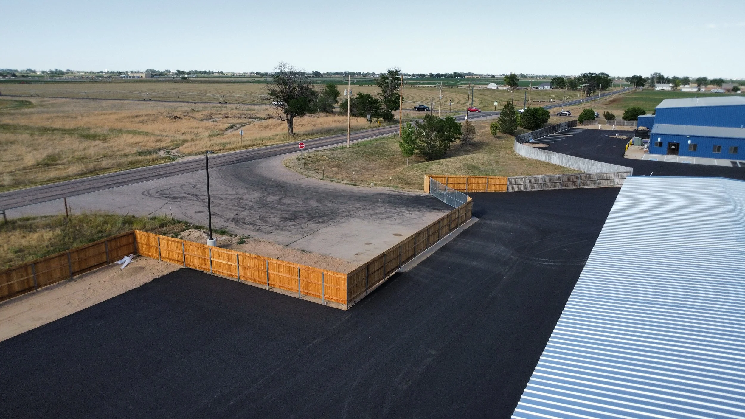 Aerial view of a parking lot and road surrounded by open fields and some trees, with a blue building on the right side and a construction fence around part of the parking lot.