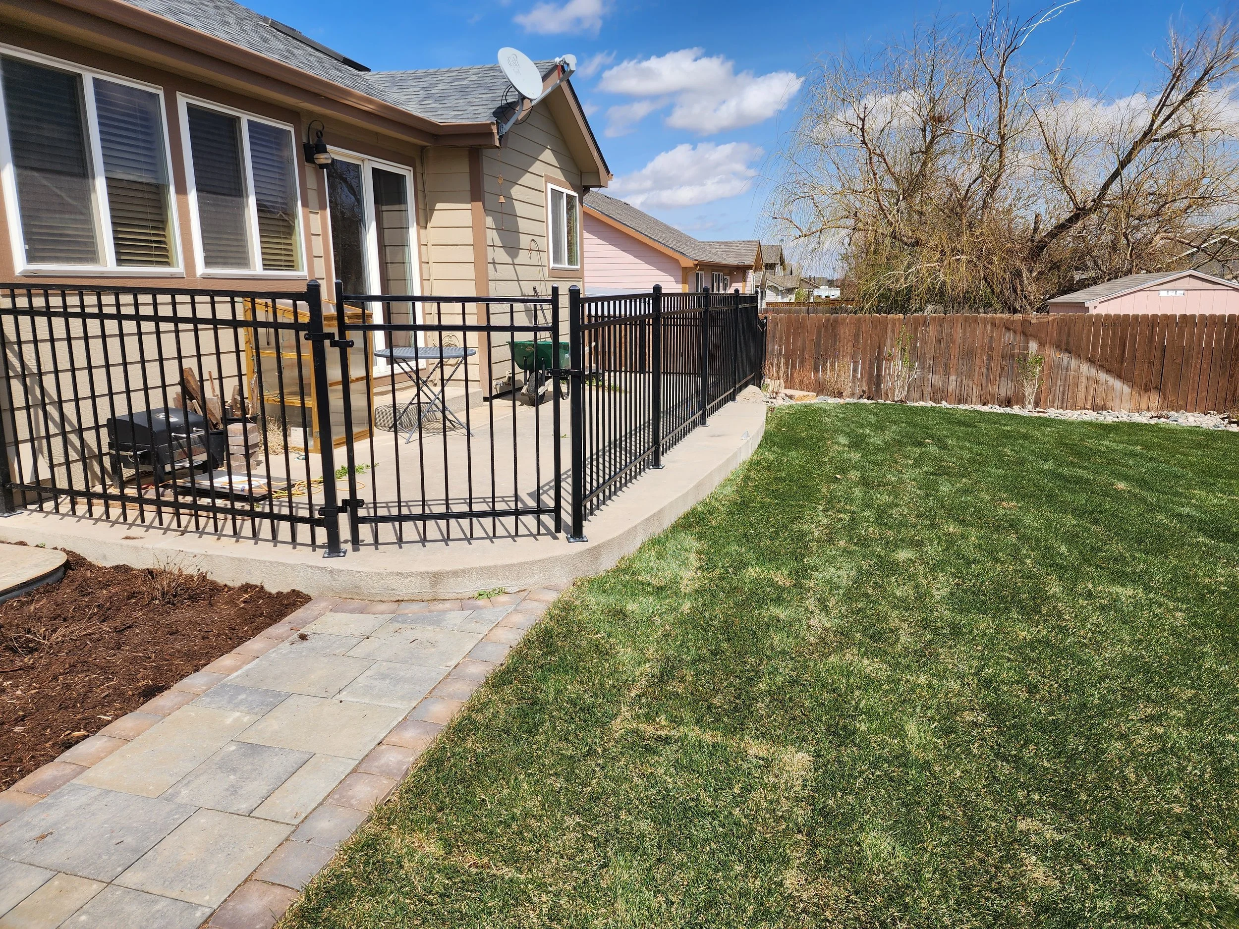 Backyard with a concrete patio enclosed by a black metal fence, a lush green lawn, a large leafless tree, and a wooden privacy fence under a blue sky with scattered clouds.