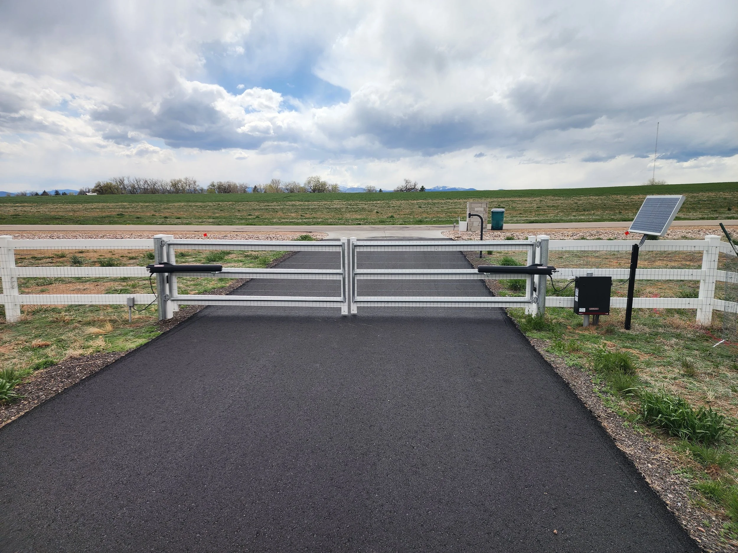 A paved driveway with a white fence on both sides leading to a gate equipped with a solar panel, outdoors in a rural area with green fields and cloudy sky in the background.