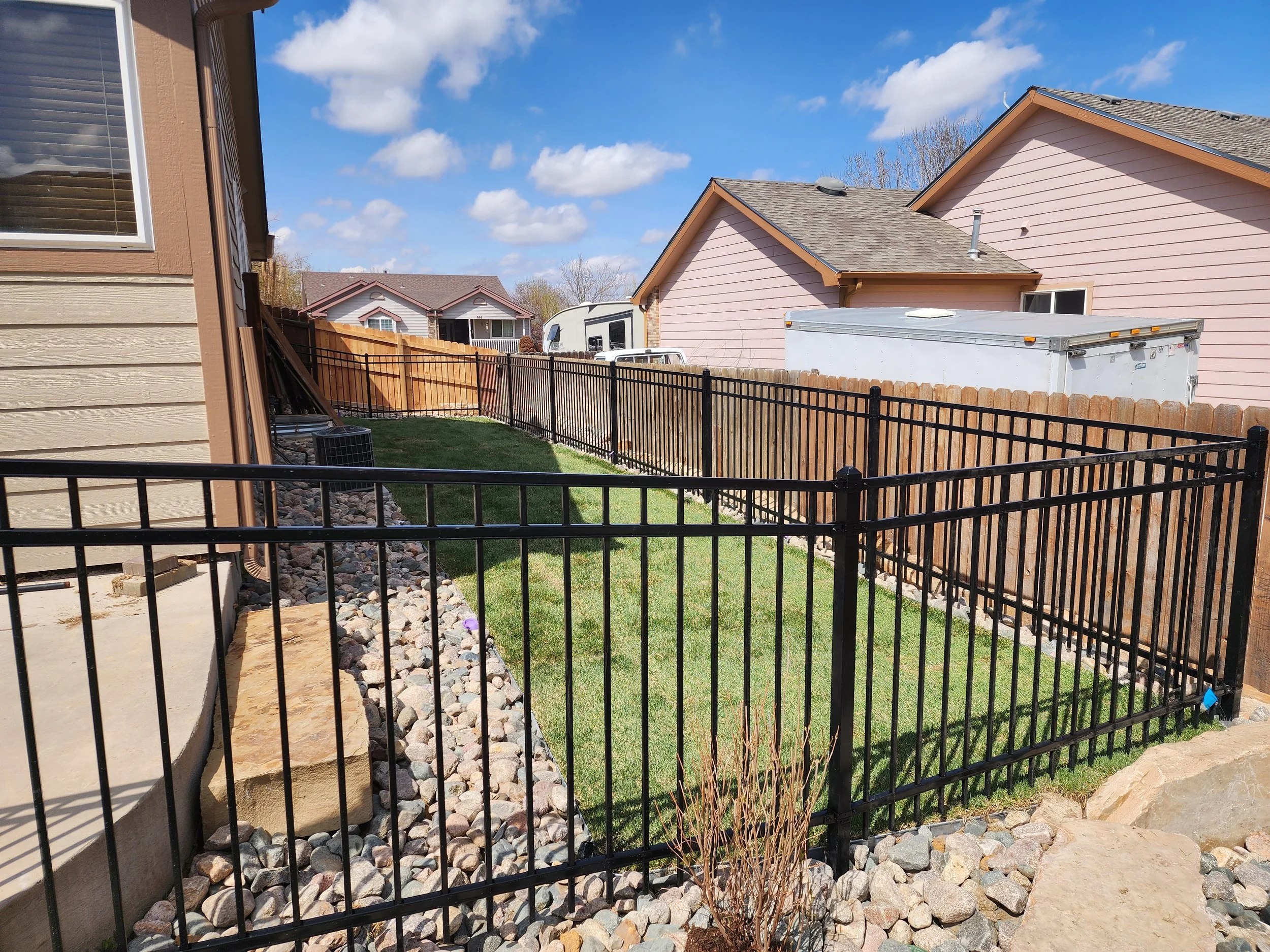 Backyard with a black metal fence, green grass, rocks, and neighboring houses with pink and beige siding under blue sky with clouds.
