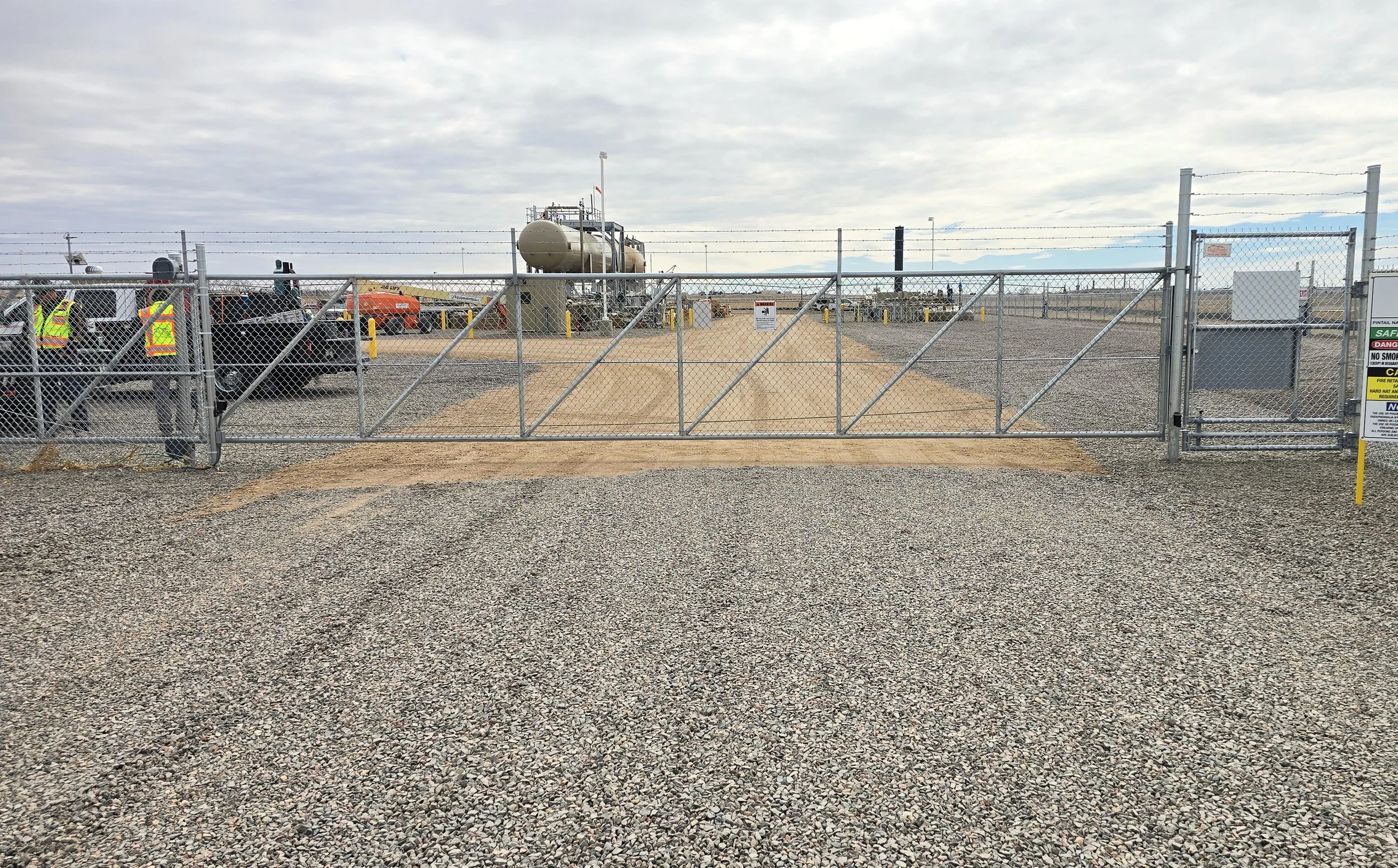 View of an industrial site enclosed by a chain-link fence with an automatic gate, various equipment, vehicles, and cloudy sky