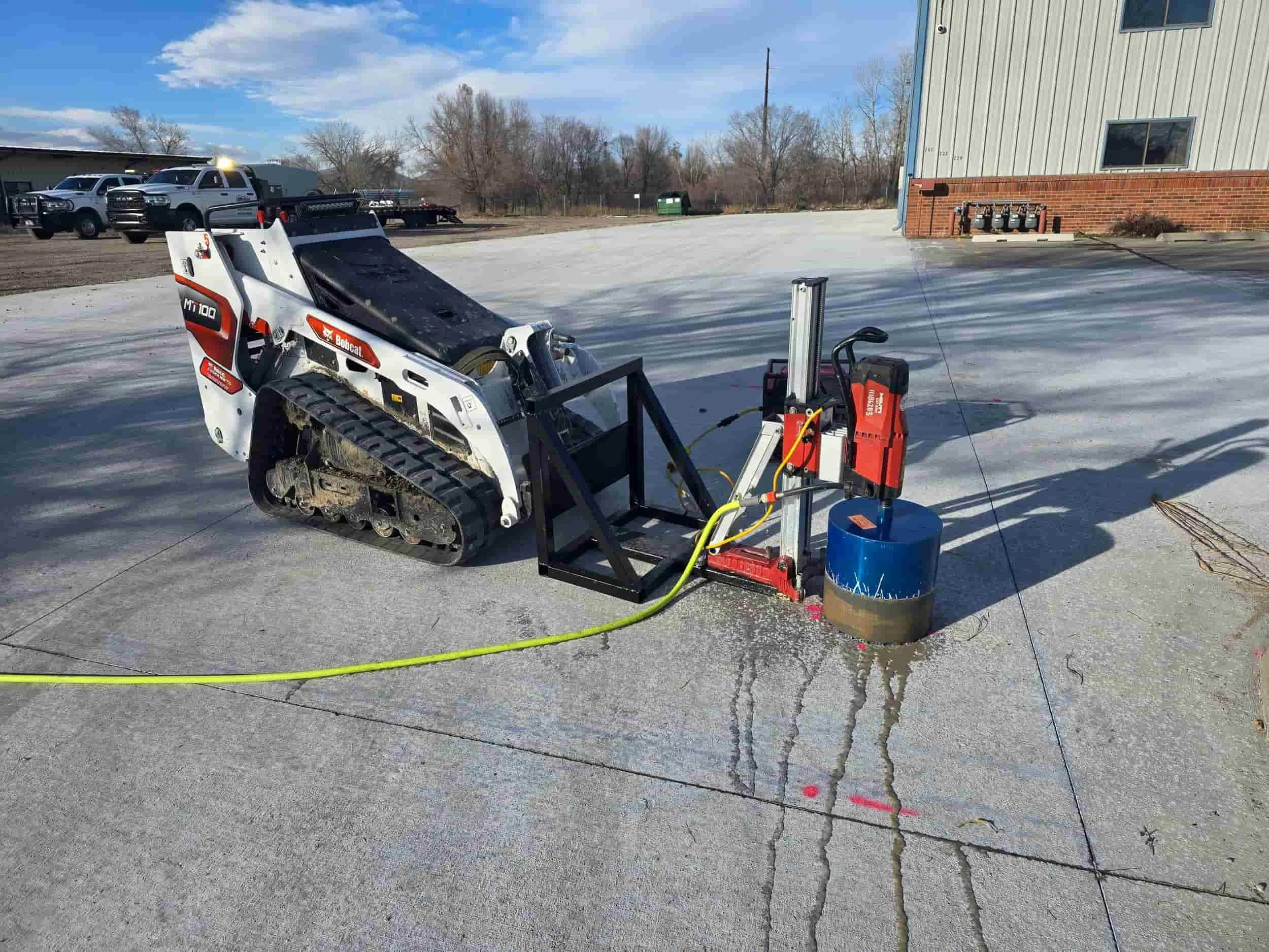 A small Bobcat tracked loader connected to a core drilling machine on a concrete surface outdoors with some trucks and a building in the background.