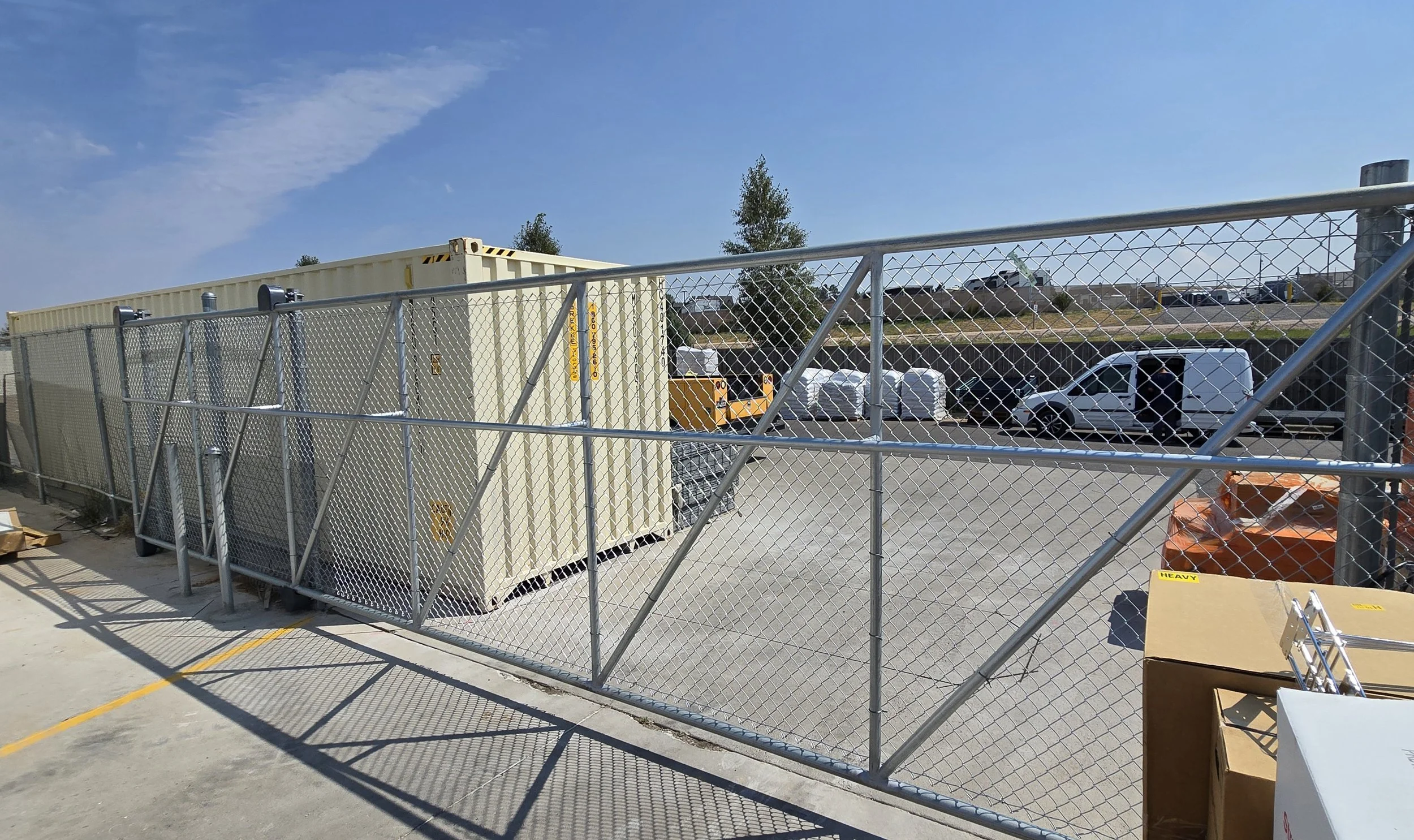 Construction site with chain-link fencing, a beige container, parked vehicles, and a blue sky with some clouds.