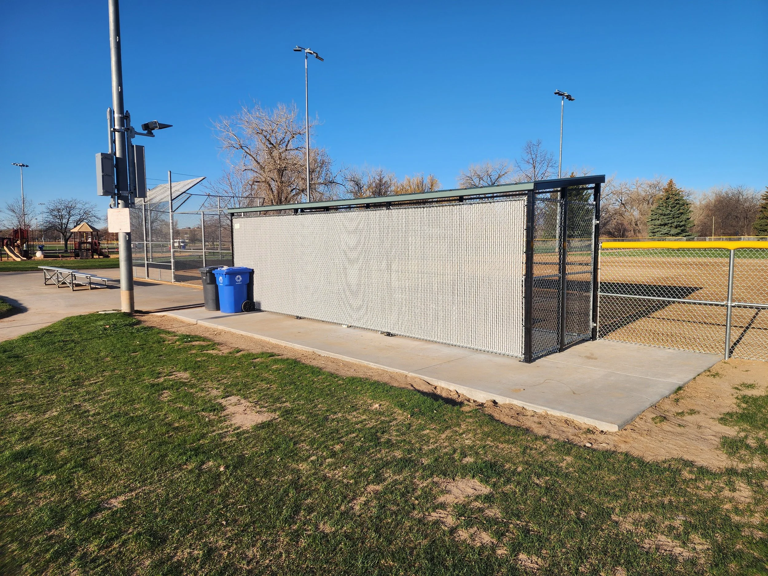 Baseball field dugout with chain-link fence and trash bins, park area with playground, trees, and clear blue sky.