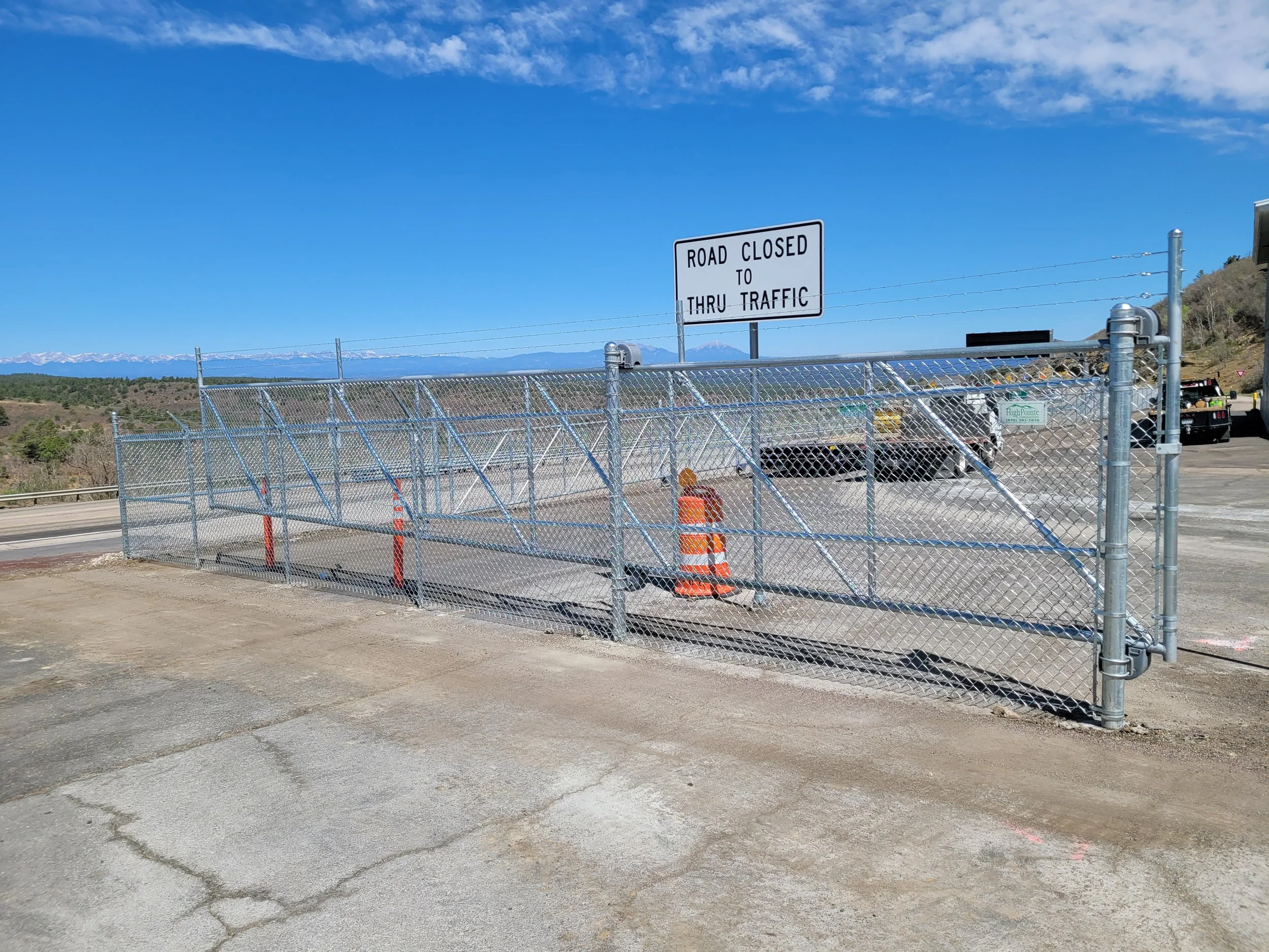 A metal chain-link fence blocking a road with a sign reading 'Road Closed to Thru Traffic' in front of a mountainous landscape under a blue sky with some scattered clouds.