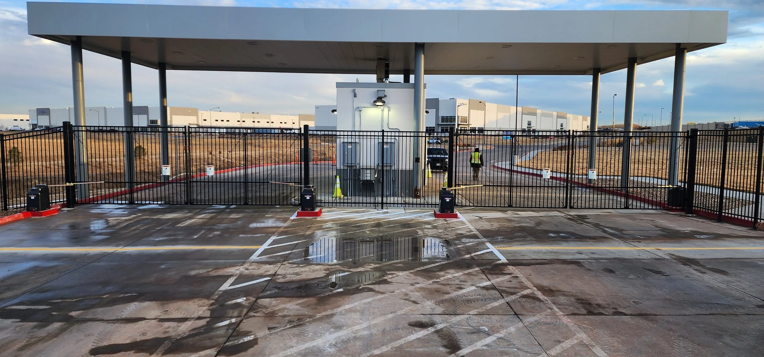 A parking lot with a gated entrance, security booth, and two workers in safety vests. The lot appears to be newly paved with some water puddles, and there are industrial buildings in the background.