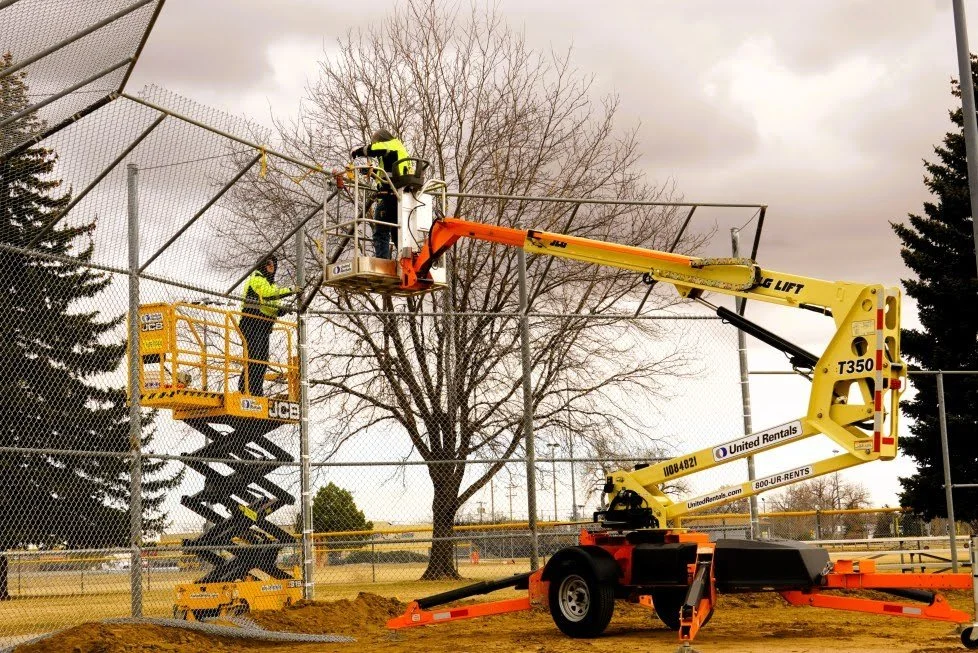 Workers in safety gear using a cherry picker lift to work on a chain-link fence at a sports field or park, with a large leafless tree and cloudy sky in the background.