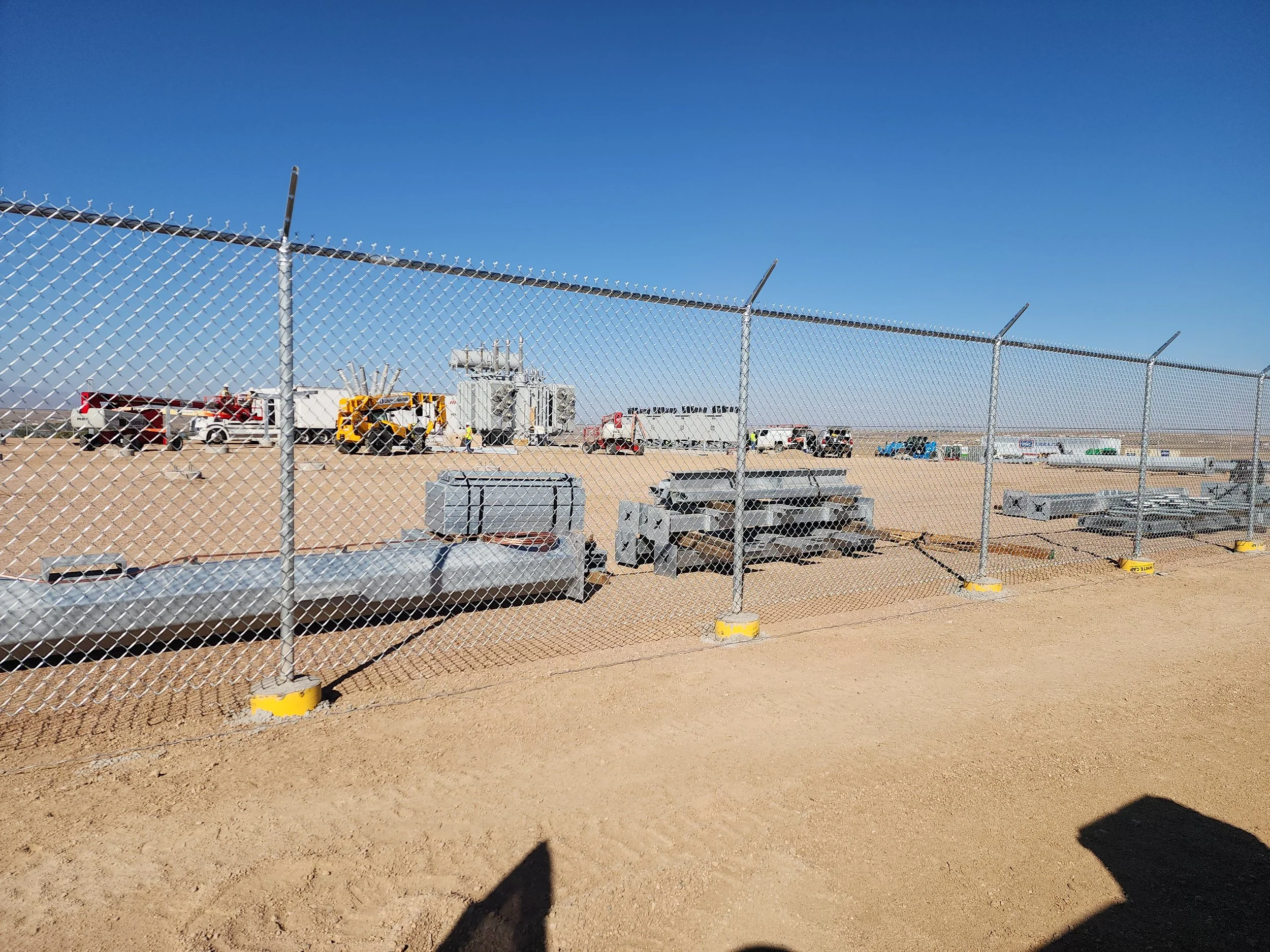 Construction site with equipment, trucks, and machinery behind a chain-link fence on a dirt ground under a clear blue sky.