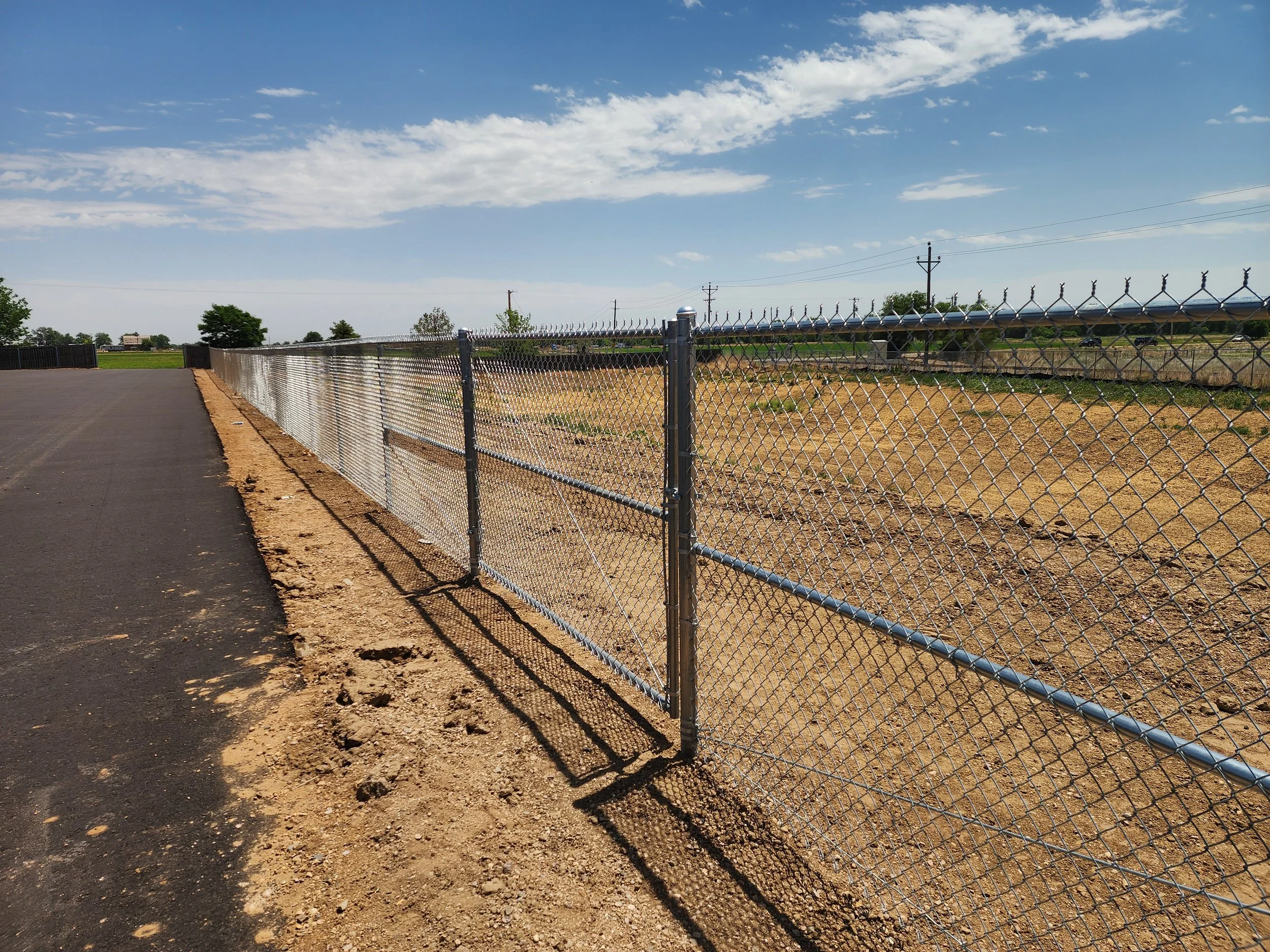 A chain-link fence runs alongside a paved road with a dirt shoulder, with a grassy field and several trees in the background. Utility poles and power lines stretch across the sky, which is mostly clear with some scattered clouds.