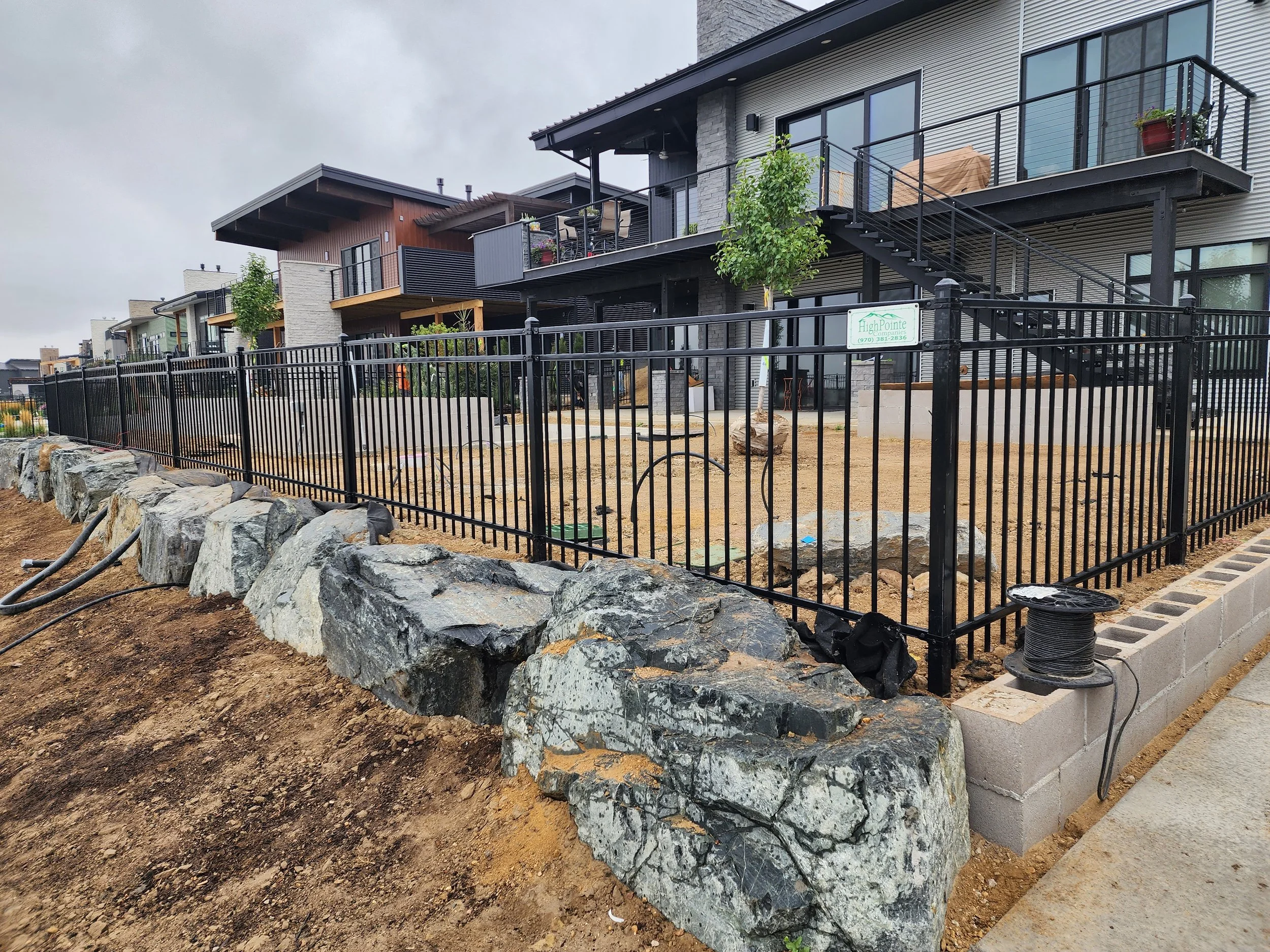 Newly installed black metal fence around a residential backyard, with large rocks along the base and a concrete block retaining wall, in front of modern multi-story homes with outdoor balconies.
