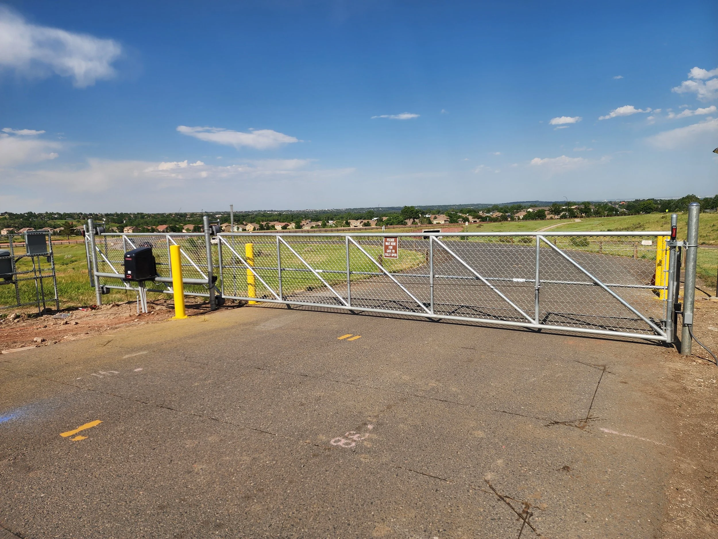 View of a highway gate with a road and suburban neighborhood in the background under a blue sky with clouds.