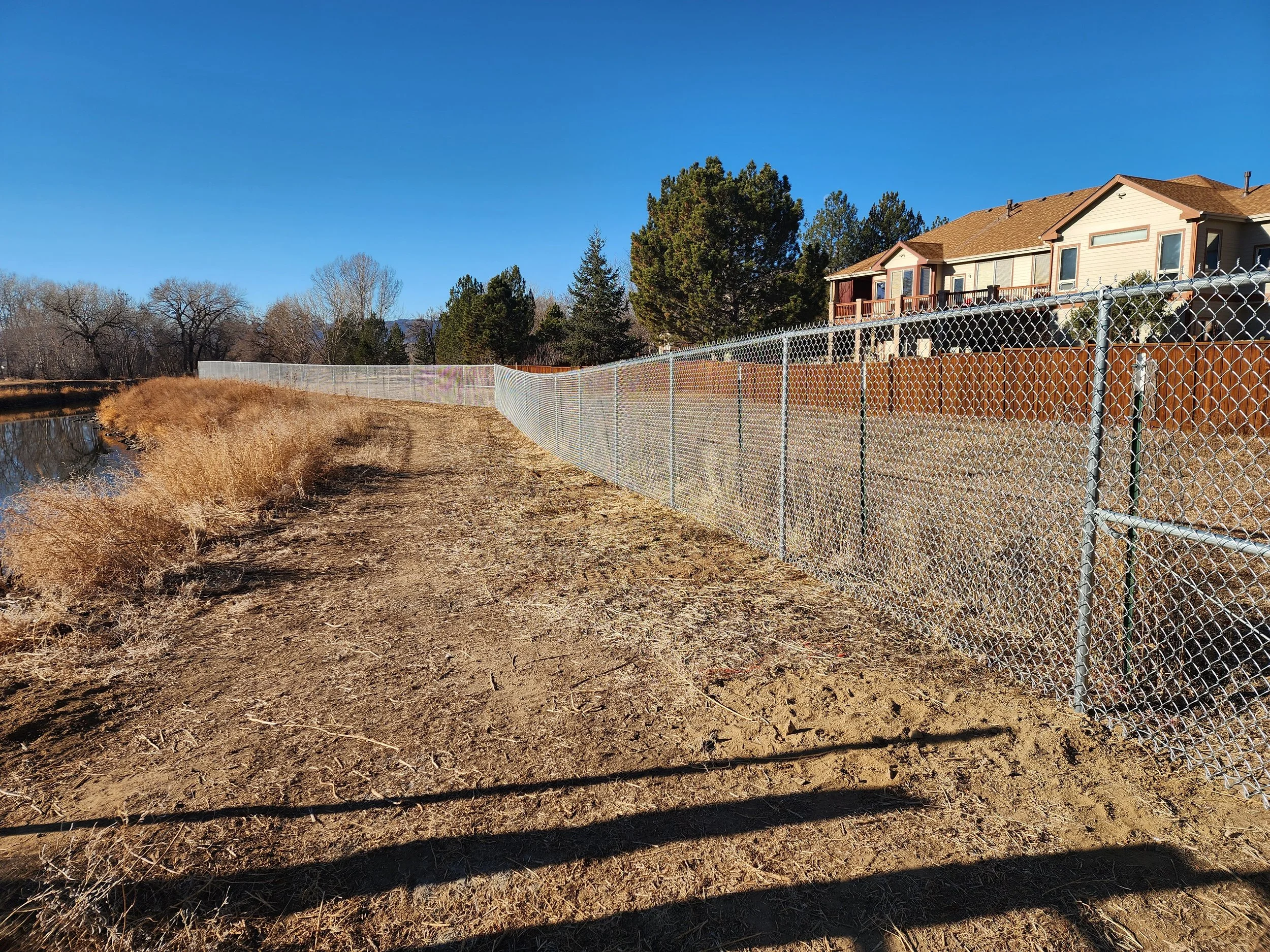 A dirt path beside a chain-link fence with houses and trees in the background on a clear day.