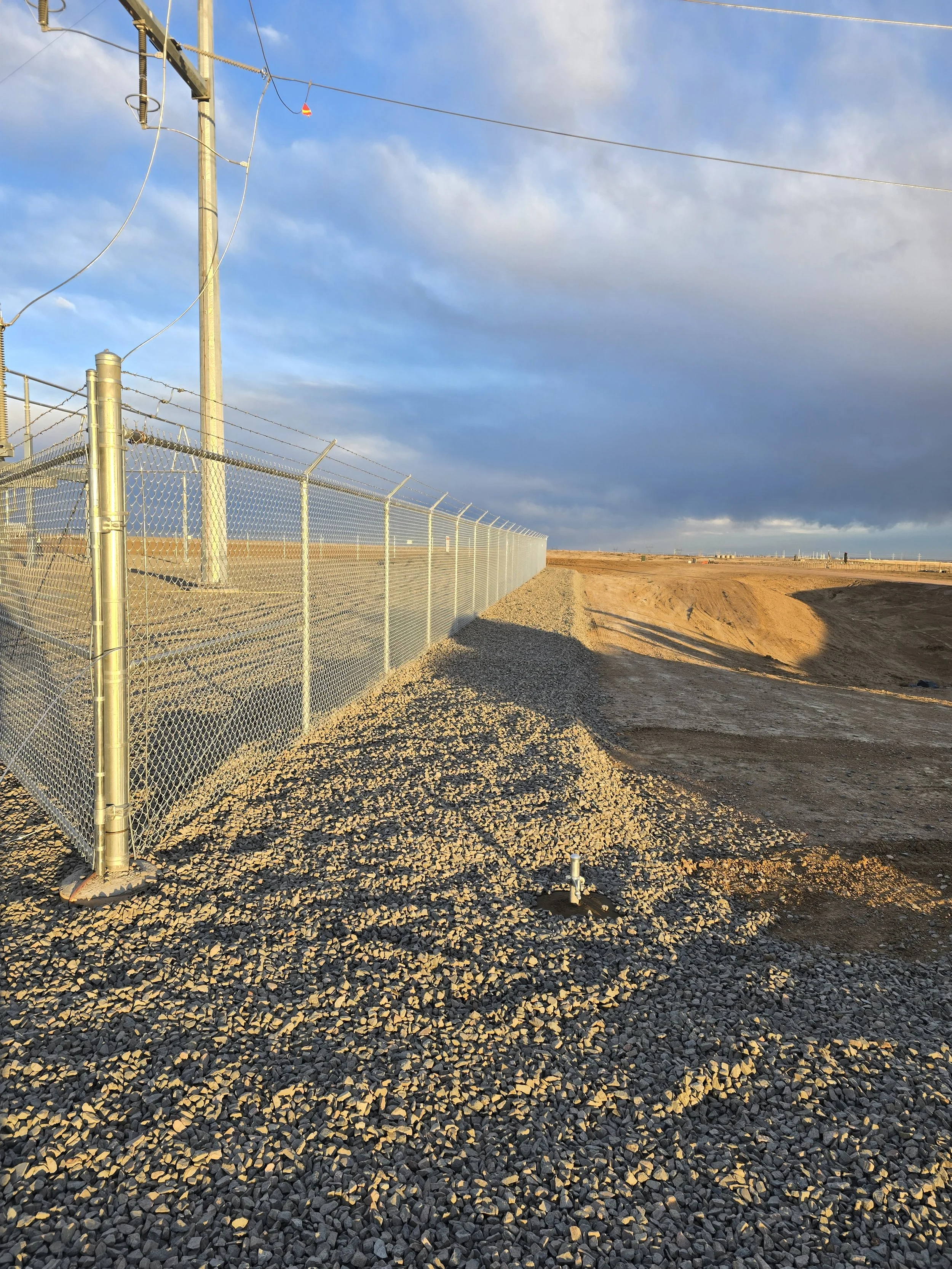 A chain-link fence running along a dirt path in a barren landscape under a partly cloudy sky at sunset.