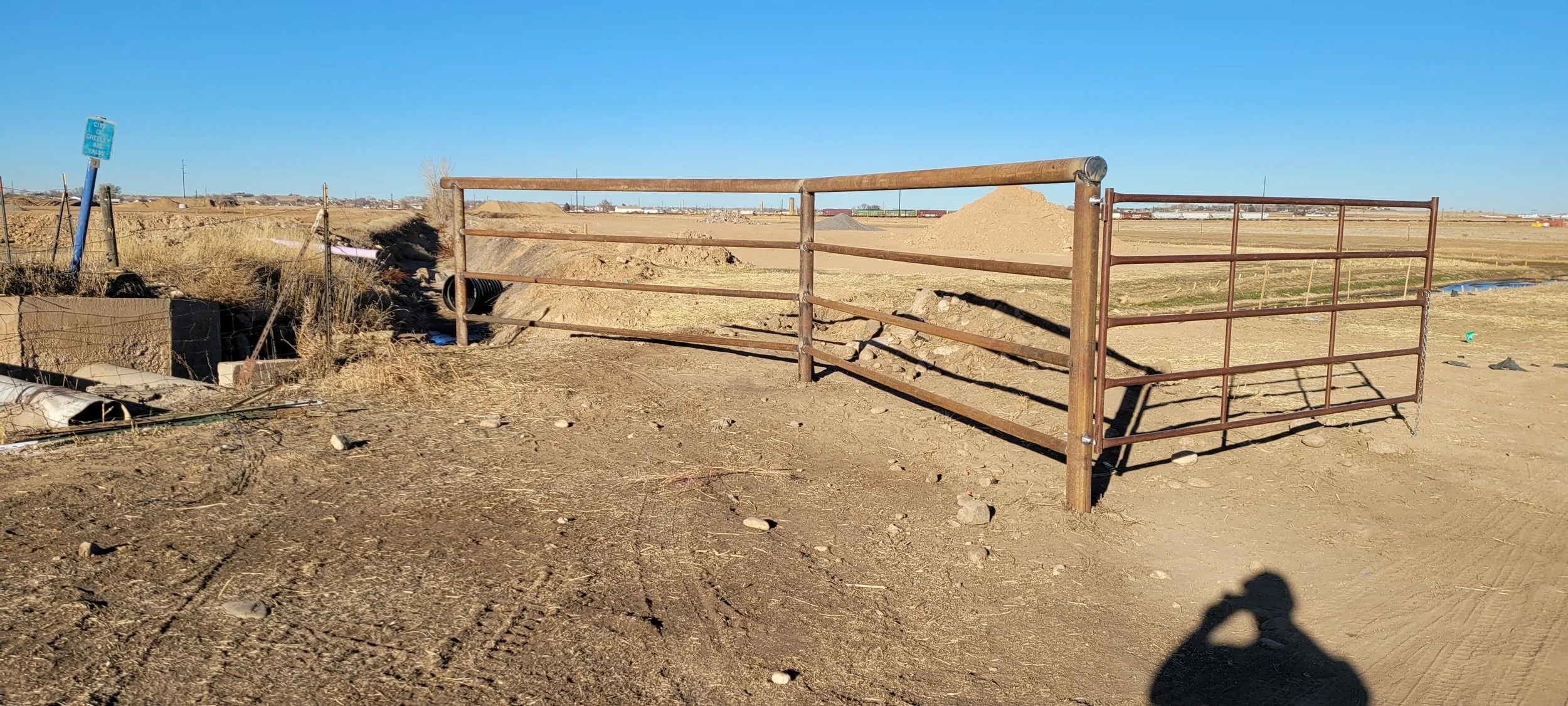 A metal horse corral gate on a dirt lot in a rural area with open fields, a bright blue sky, and distant farm structures.