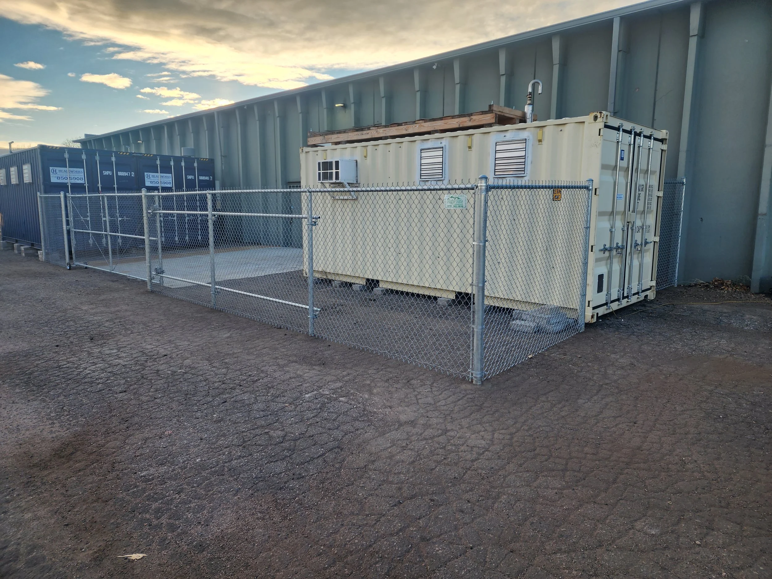 A fenced outdoor area with a beige shipping container, an air conditioning unit, and a metal utility box next to a large building with a metal exterior. The ground is gravel, and the sky is partly cloudy at sunset.
