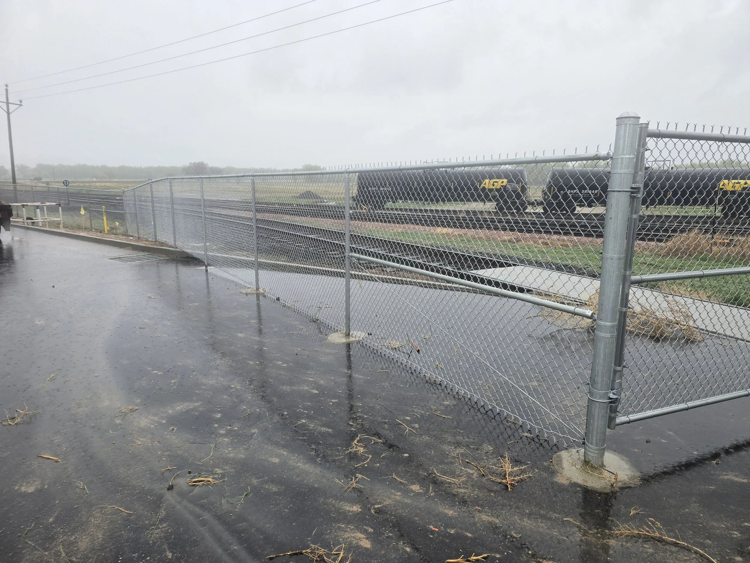Flooded parking lot with a chain-link fence and train tracks in the background during rainy weather.
