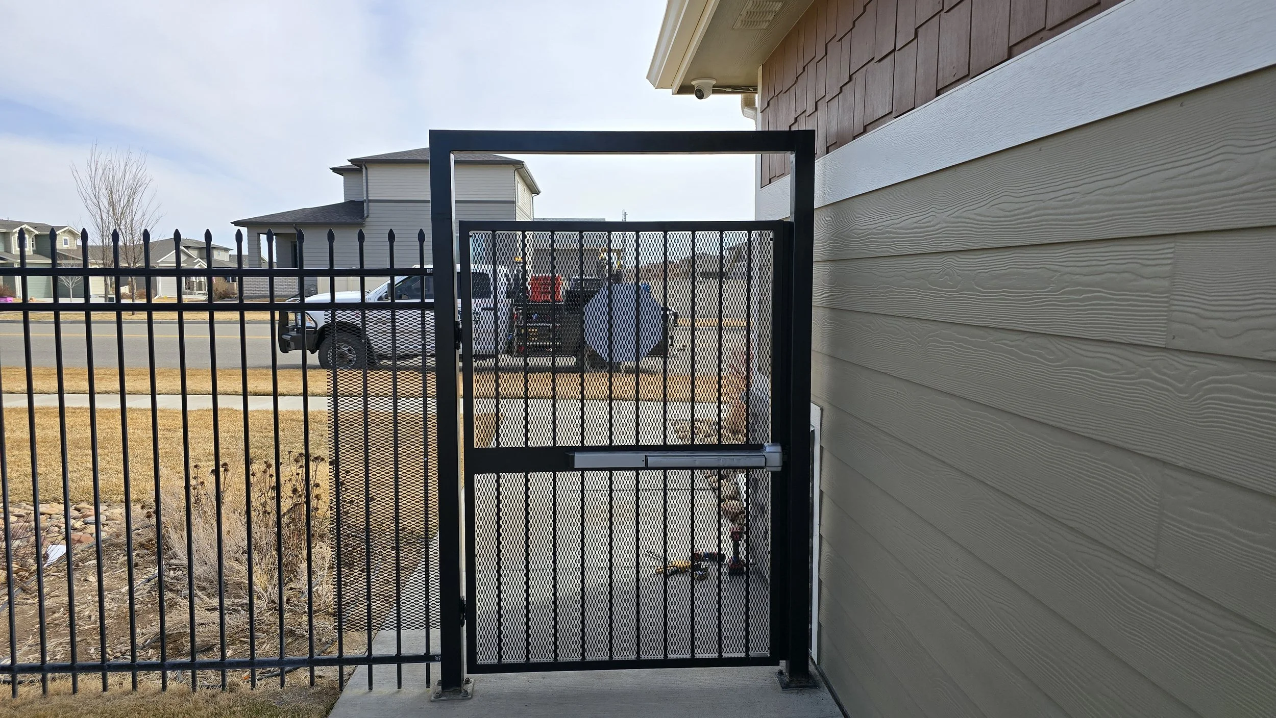 View of a black metal gate with a mesh design, attached to a beige house with vinyl siding, leading to a sidewalk and street with parked cars and apartment buildings in the background.
