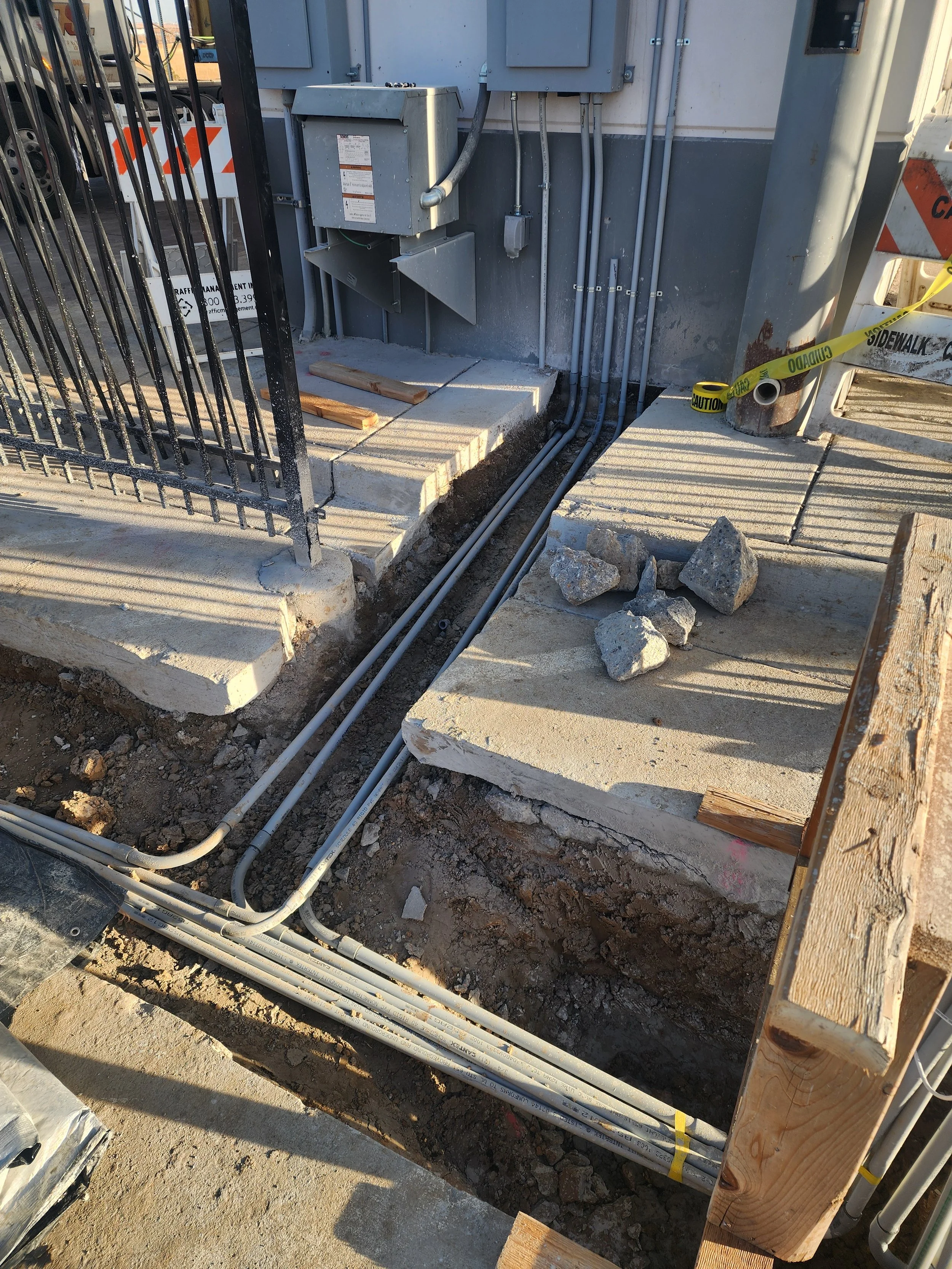 Construction site with exposed electrical and plumbing conduits, concrete slabs, rocks, caution tape, and construction barriers.