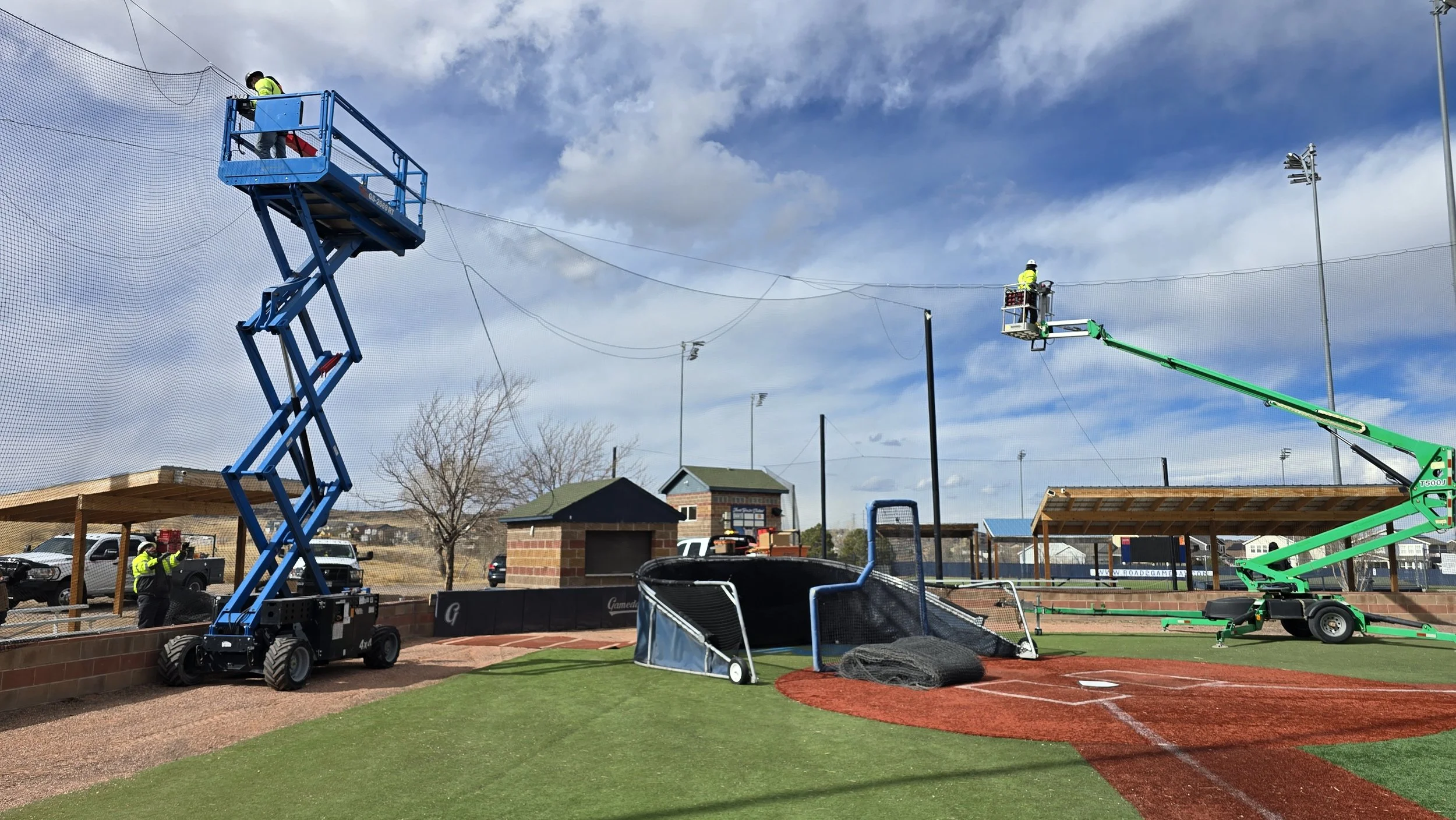 Workers use hydraulic lifts to repair or install safety nets around the sports field at a stadium, with a baseball diamond visible in the foreground.