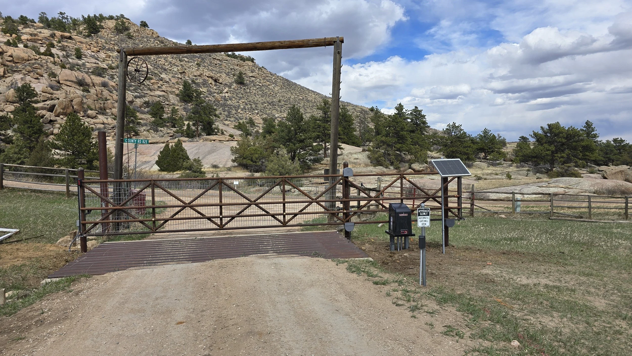 A fenced entrance gate made of wooden posts and metal fence panels on a dirt road, with a small solar panel on a stand, nearby mailboxes, and a sign indicating approaching slowly for automatic gate access, with trees, rocky hills, and a partly cloudy