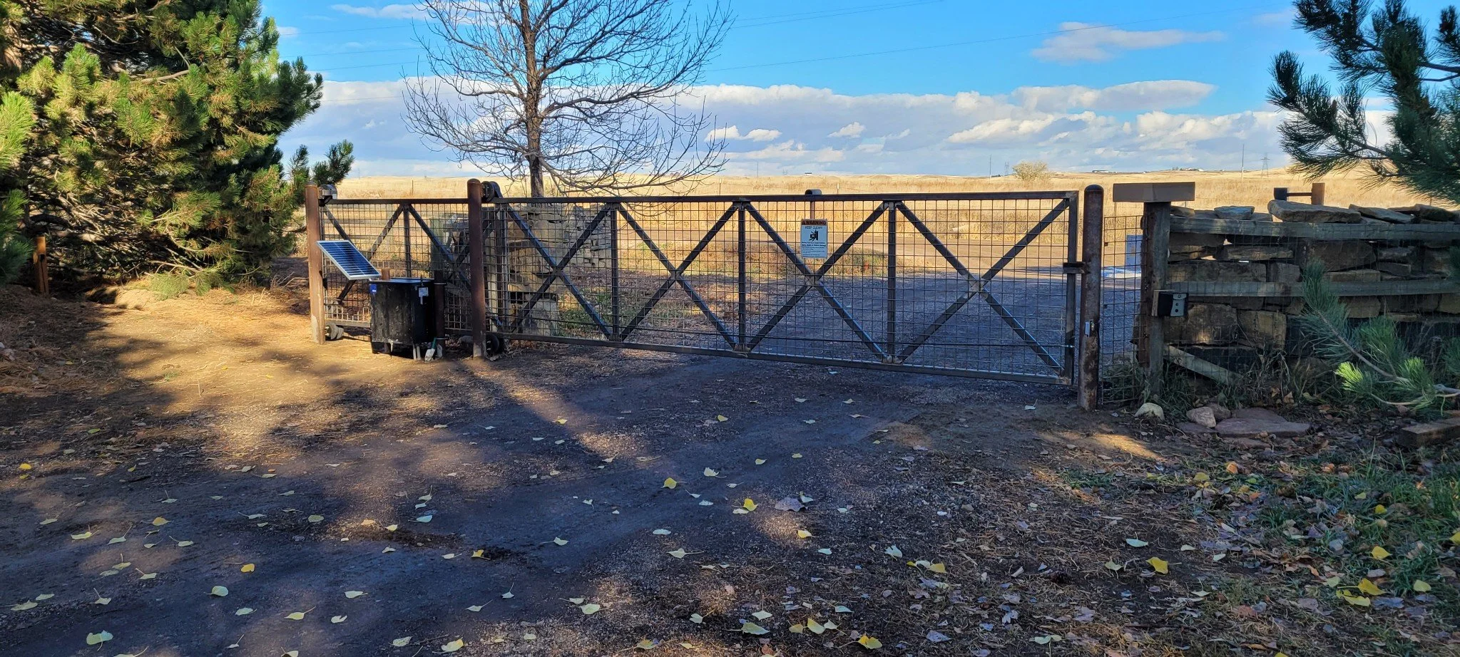 A metal gate with a sign, surrounded by trees and a dirt ground with fallen leaves, leading to a wide open field under a partly cloudy sky.