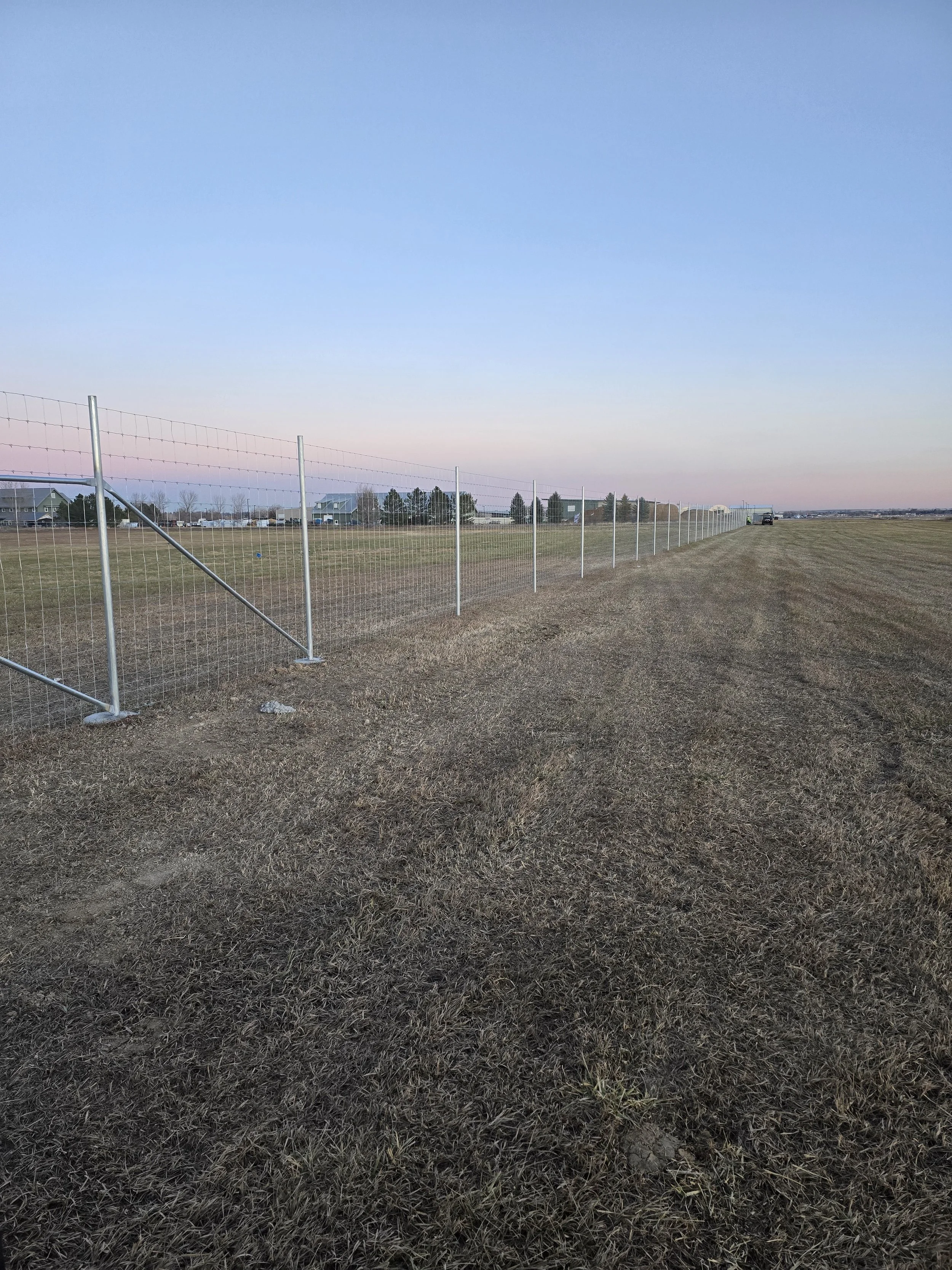 An open field with a wire fence running diagonally across the foreground, houses in the distance, and a car parked far to the right under a clear sky.