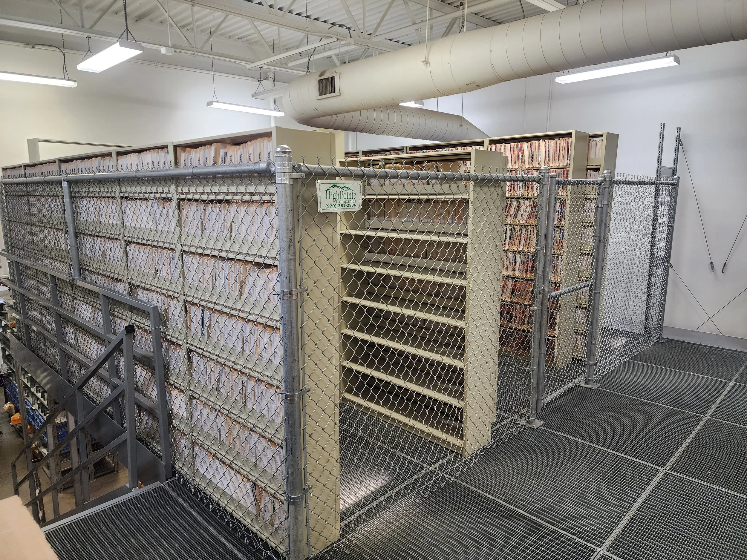 A storage room with metal shelving filled with files, enclosed by a chain-link fence with a sign, in an industrial or warehouse setting.