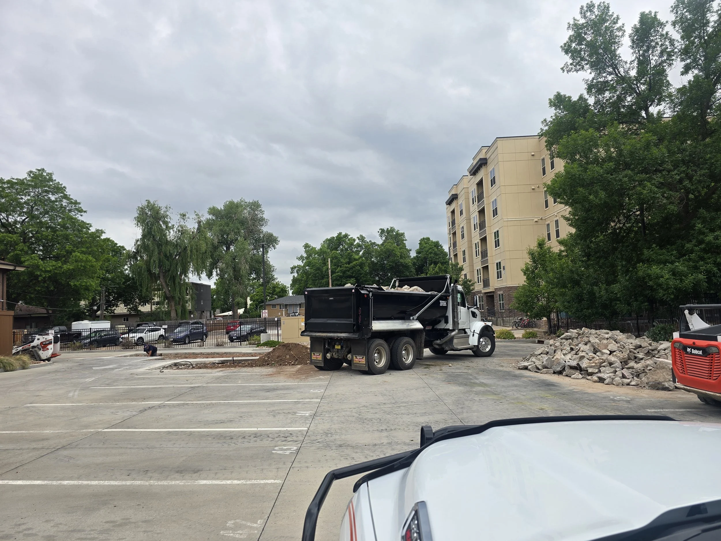 Construction site in a parking lot with a dump truck, rocks, and construction worker, surrounded by trees and apartment buildings on a cloudy day.