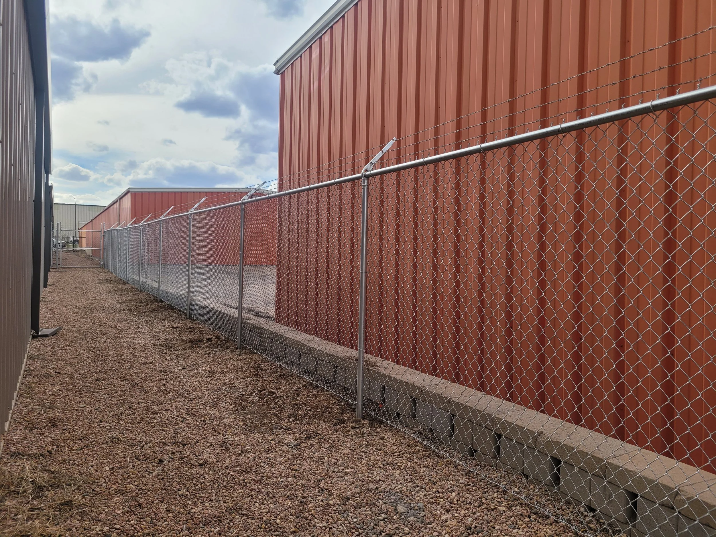 Photo of a chain-link fence running along the side of a building with red and gray metal walls, gravel ground, and a cloudy sky overhead.