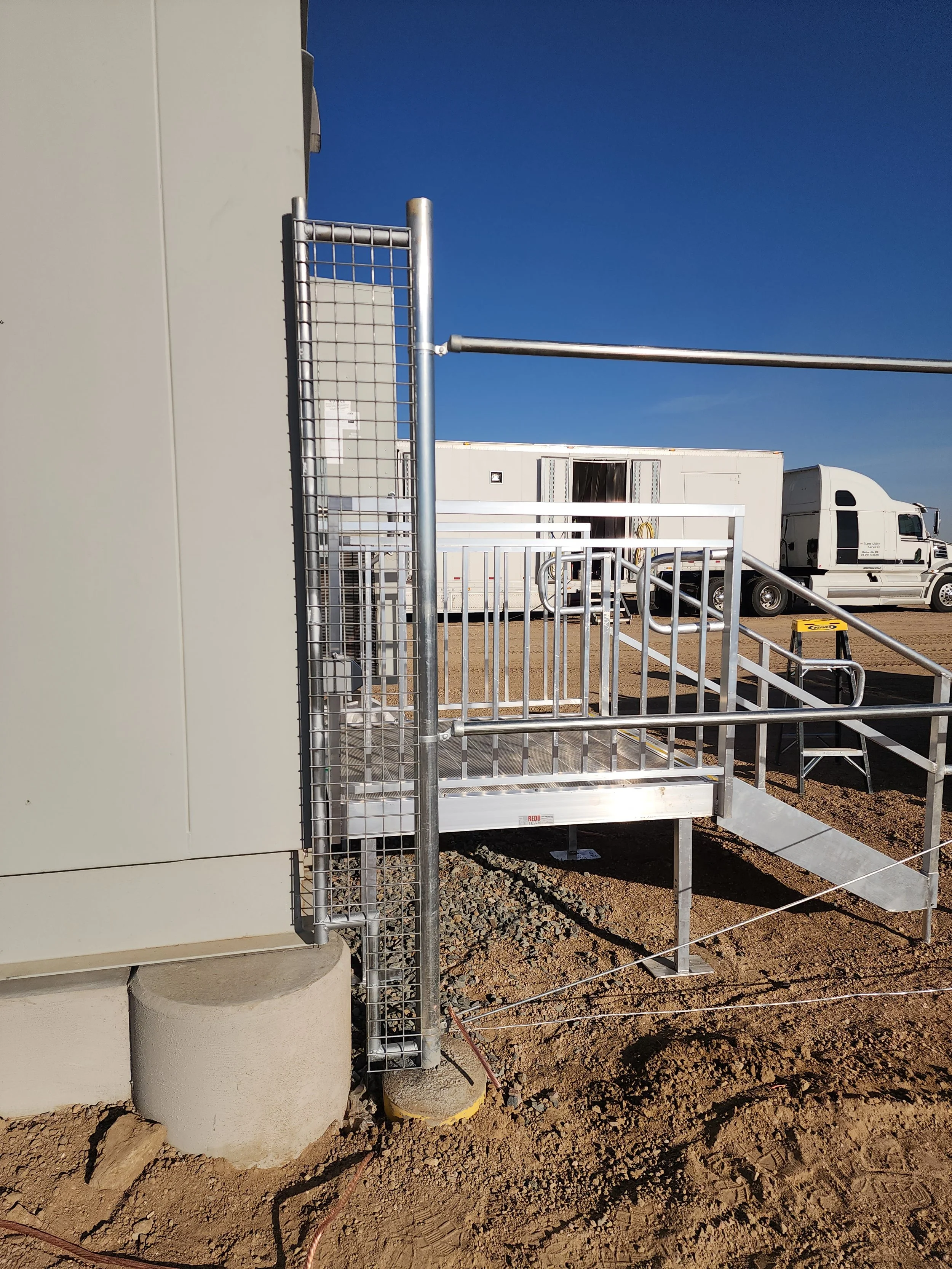 Metal wheelchair ramp with handrails leading to a building entrance, with a dirt ground and trucks parked in the background under a clear blue sky.