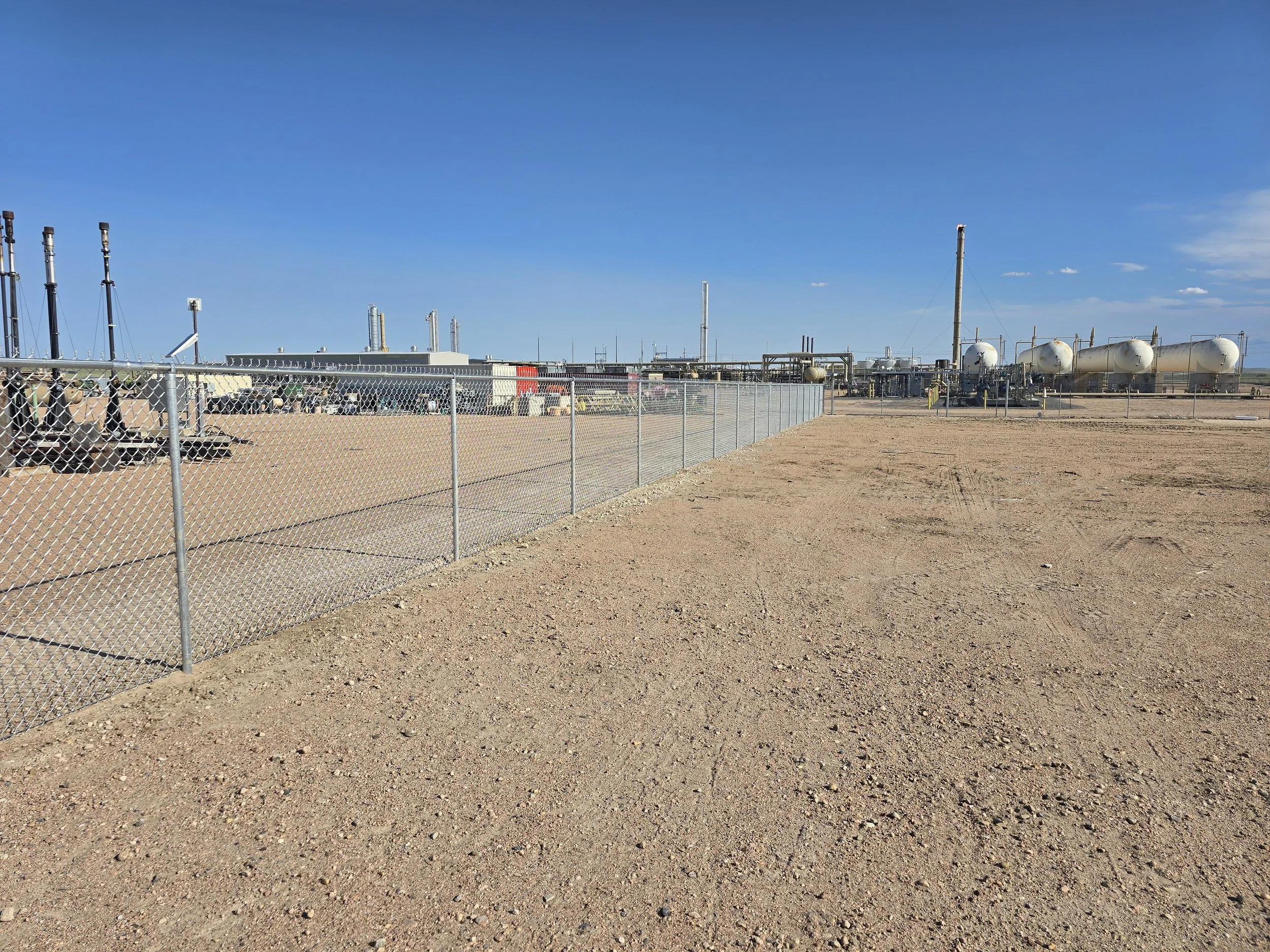 Industrial facility with storage tanks, pipelines, and equipment behind a chain-link fence in a desert landscape under a clear blue sky.