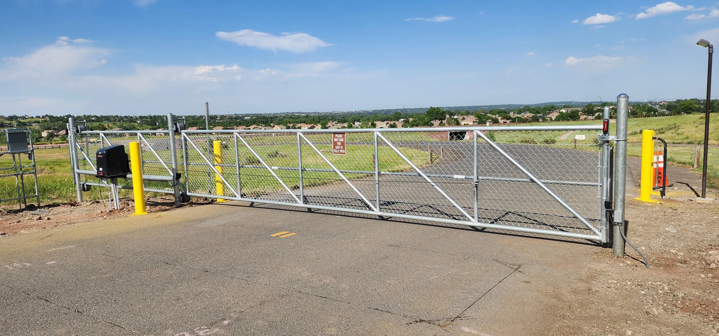 A closed chain-link gate with yellow safety posts on either side, set in a rural area with green fields and houses in the distance, under a partly cloudy sky.
