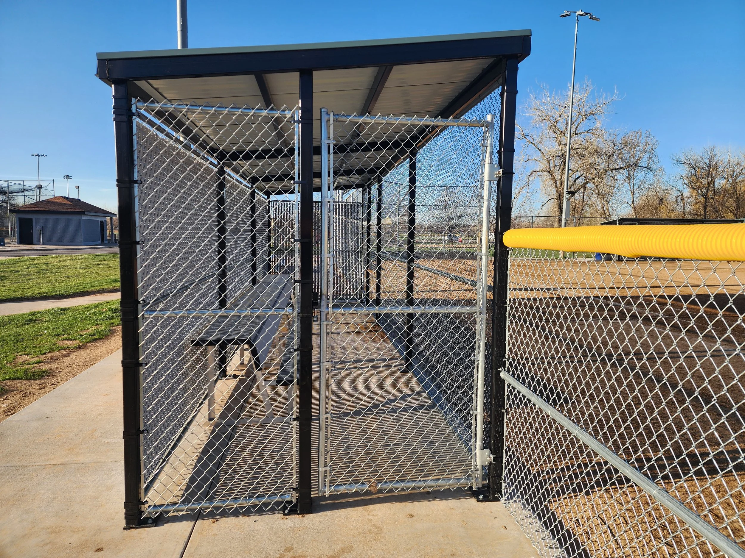 A chain-link fence enclosure with a black metal frame and a swinging gate, situated on a concrete sidewalk near a park or sports field, with a yellow protective tube on the fence on the right side, and trees and open fields in the background.