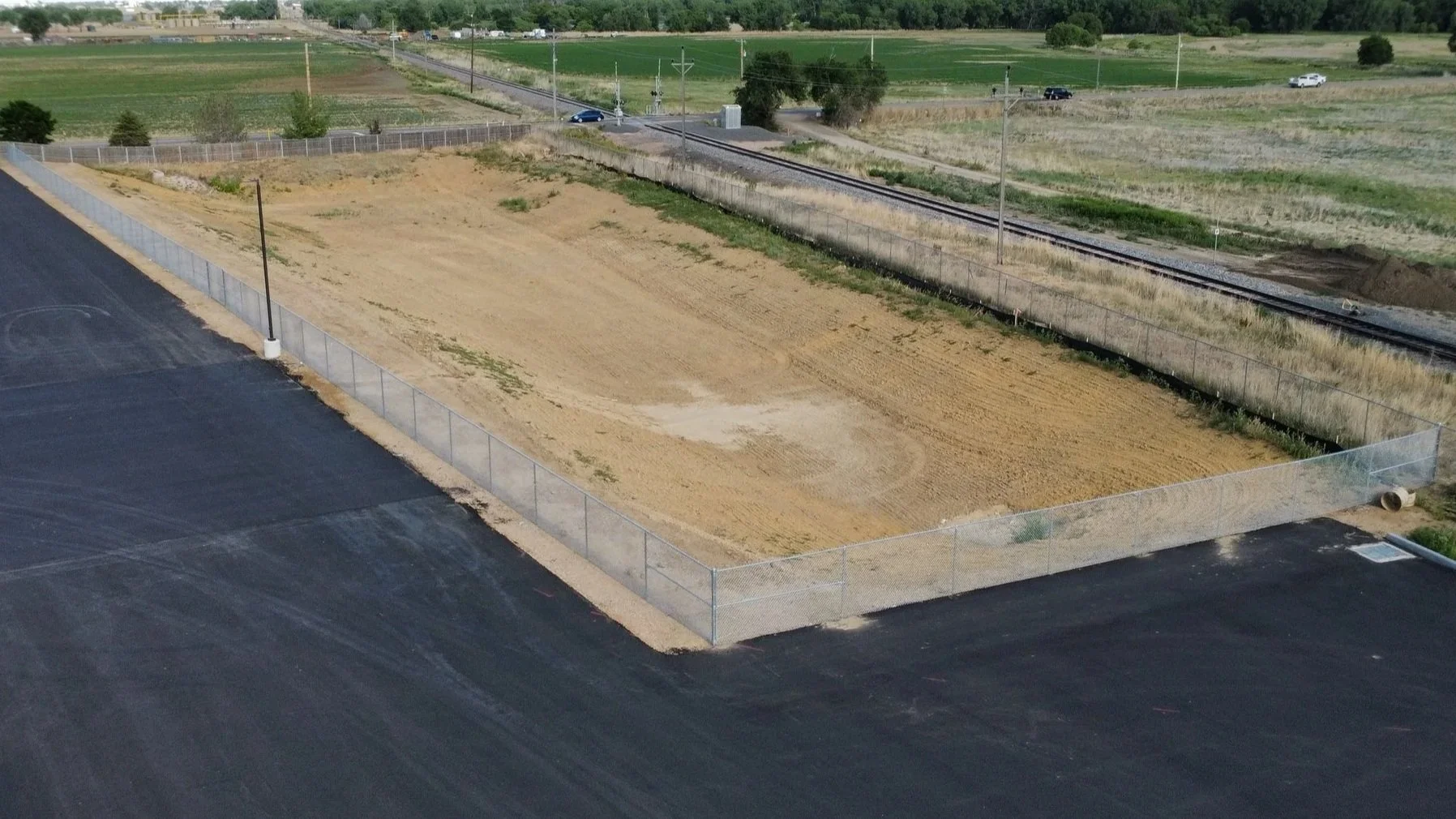 An empty construction site with a dirt lot enclosed by a chain-link fence, adjacent to a black asphalt parking lot, with train tracks and green fields in the background.