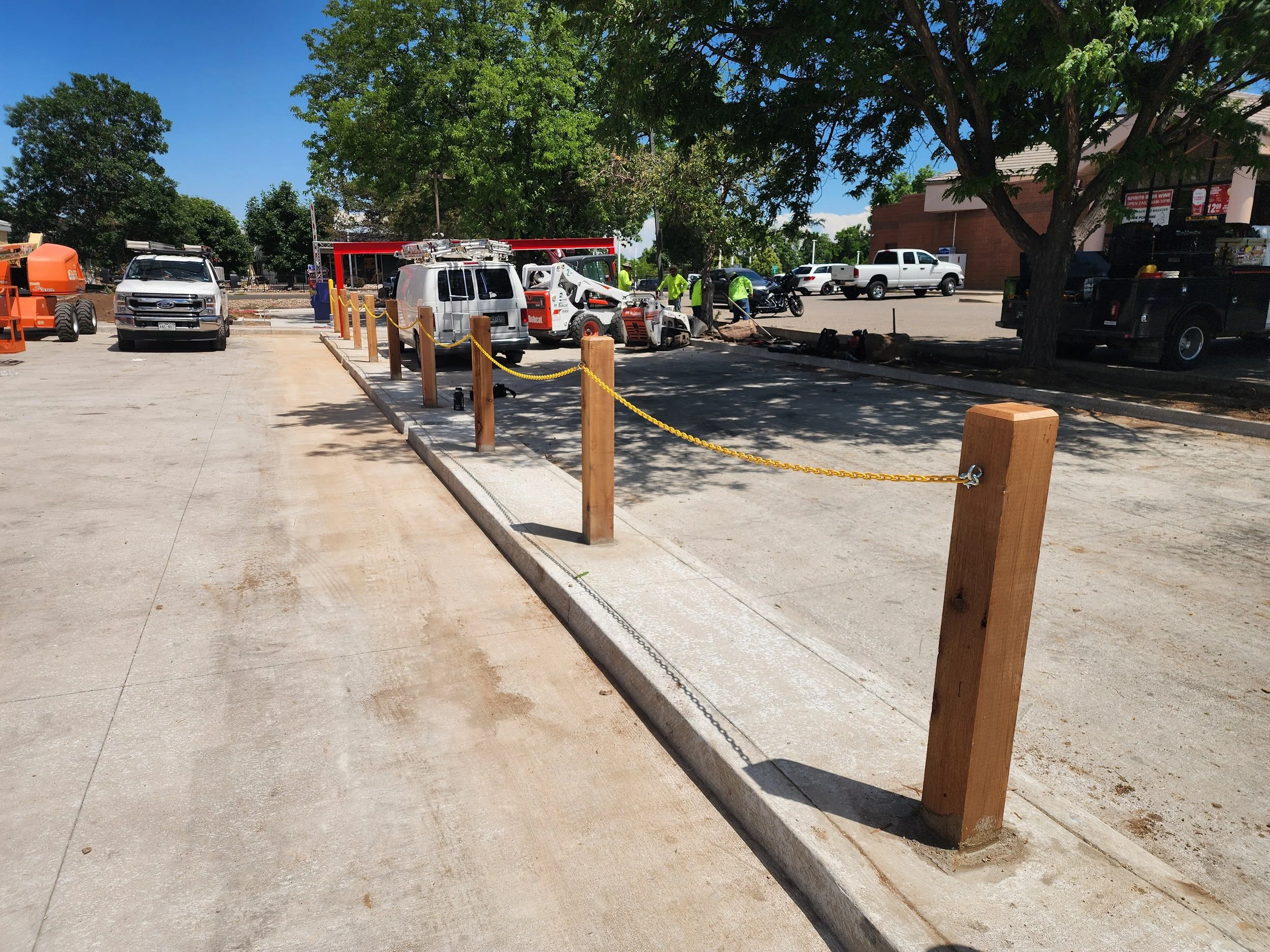 Construction site with wooden posts and yellow chain barrier, sidewalk, trees, construction vehicles, and workers in the background.