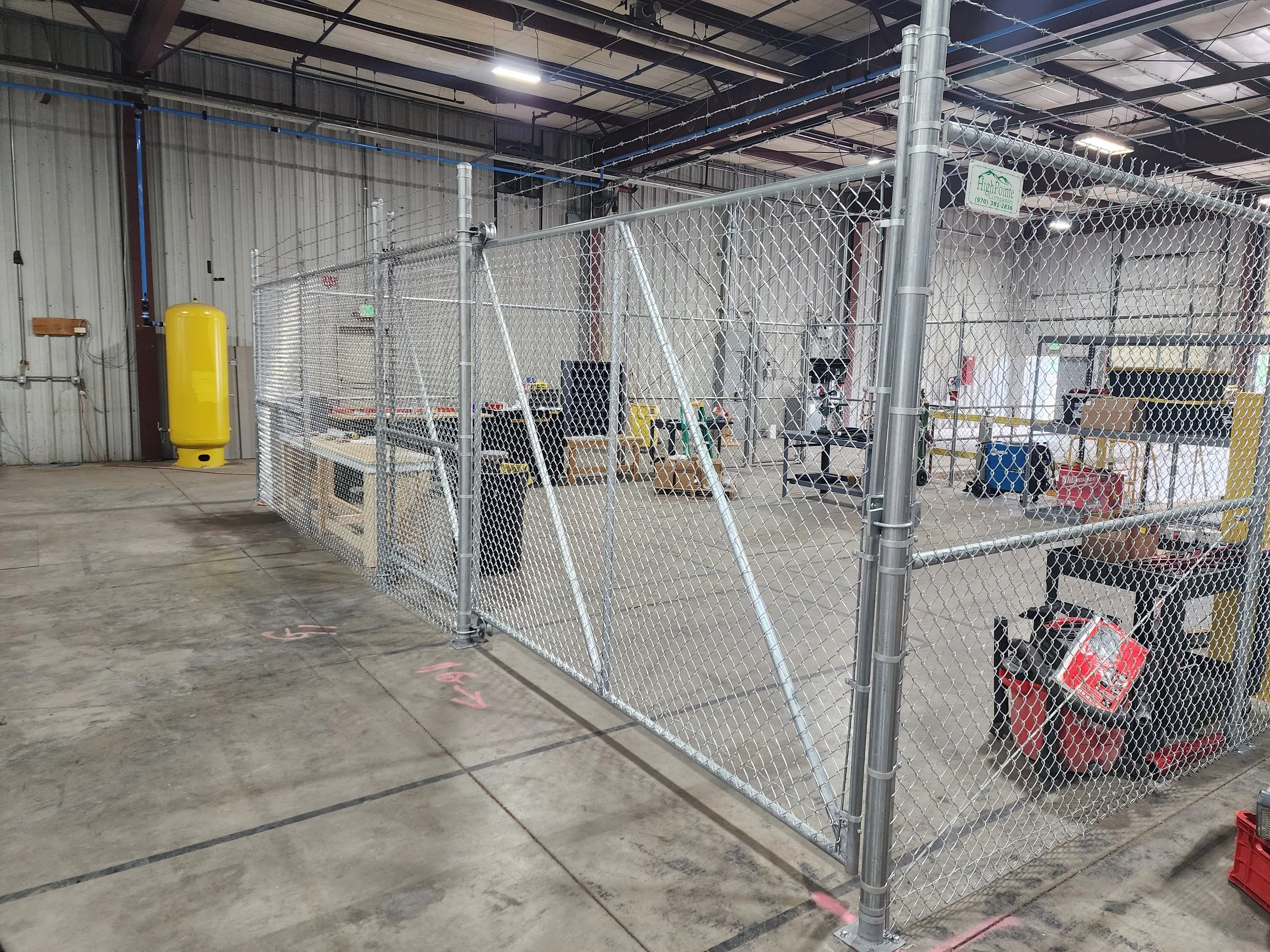 Inside a large industrial warehouse with a fenced-off work area. The area contains various tools, equipment, and supplies, including a large yellow tank, storage boxes, and workbenches. The warehouse has metal walls and a high ceiling with exposed be