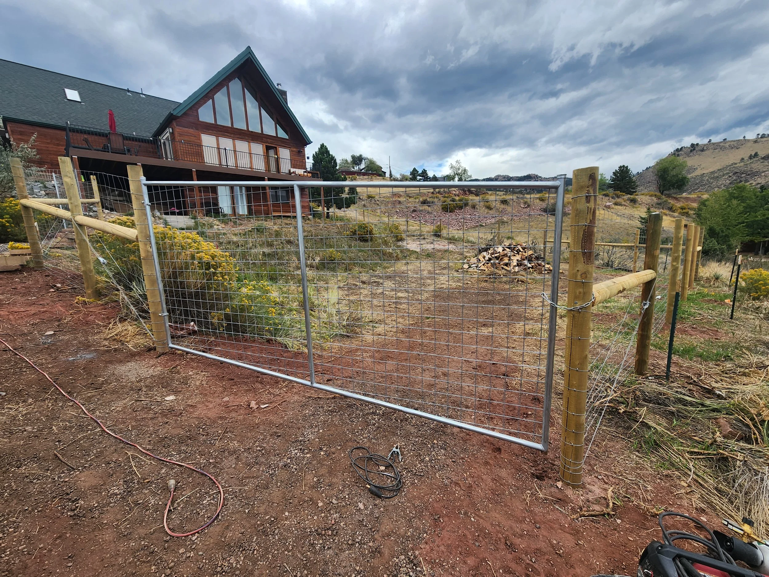 A metal gate with a lock on a dirt path, surrounding a hillside with yellow flowering bushes, and a wooden house on the hill in the background under a partly cloudy sky.