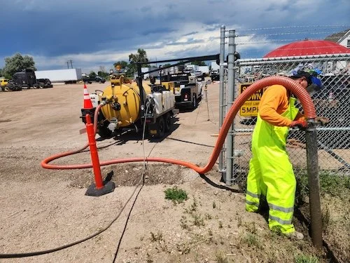 Worker in safety gear operating equipment near a chain-link fence, with a large yellow vacuum truck and construction vehicles in the background under a partly cloudy sky.