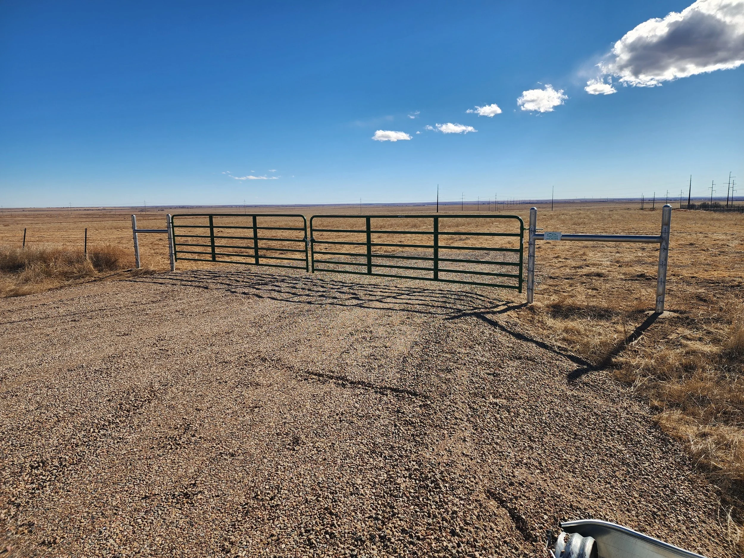 Open green metal gate on a gravel road leading into flat, expansive dry grassland under a clear blue sky with a few small clouds.