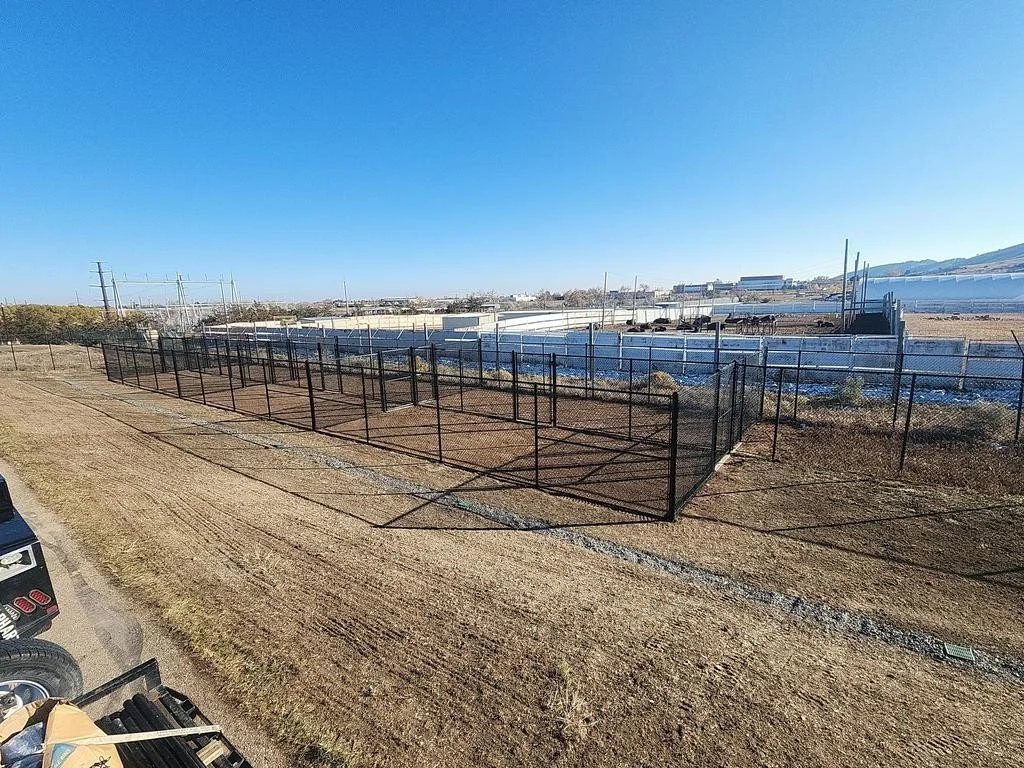 A fenced outdoor area with a dirt ground in an industrial or construction setting, with a large white building and power lines in the background.