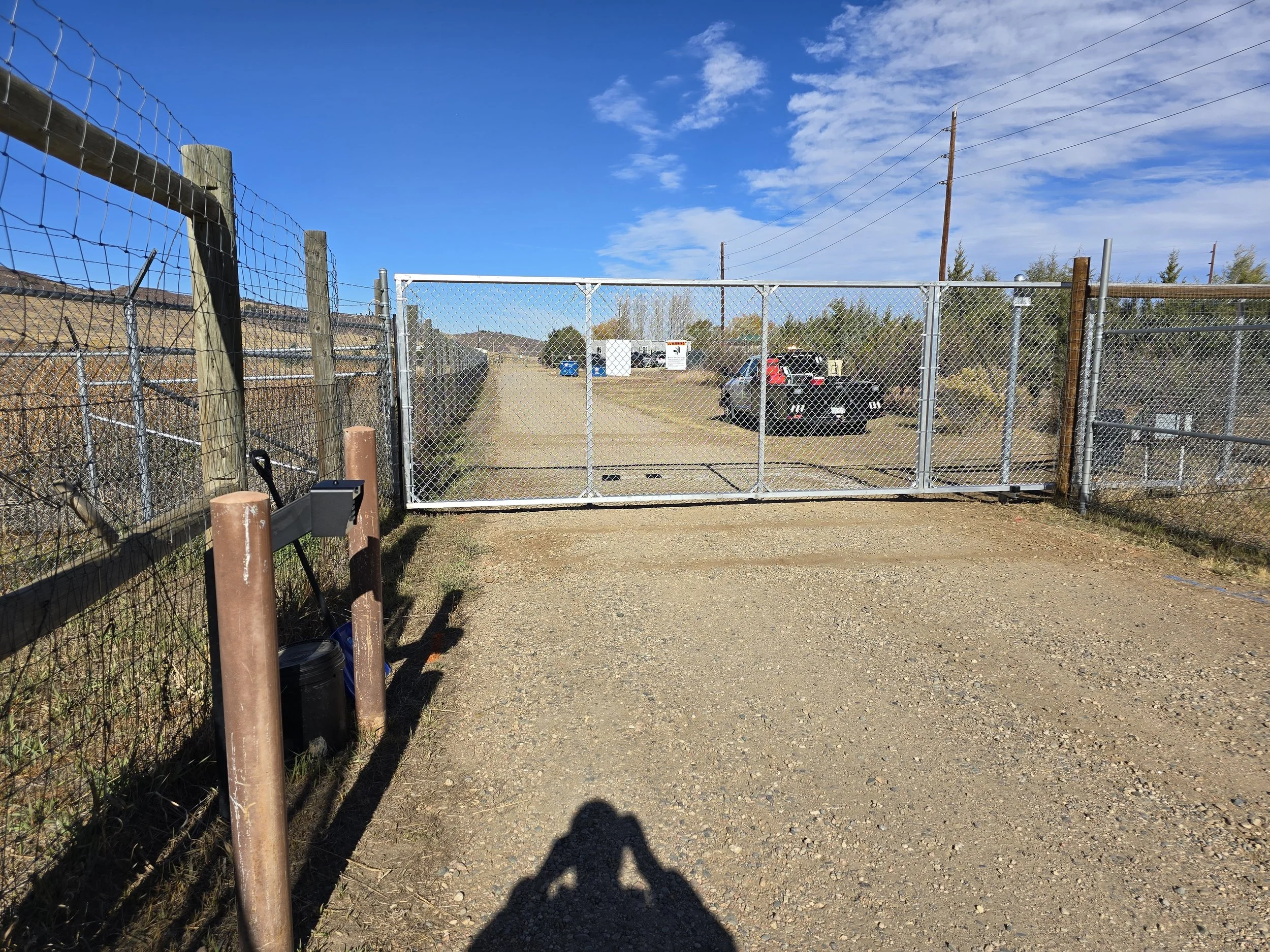 A gravel driveway with a chain-link gate blocking the entrance, surrounding a parking lot with several parked cars, under a blue sky with scattered clouds.