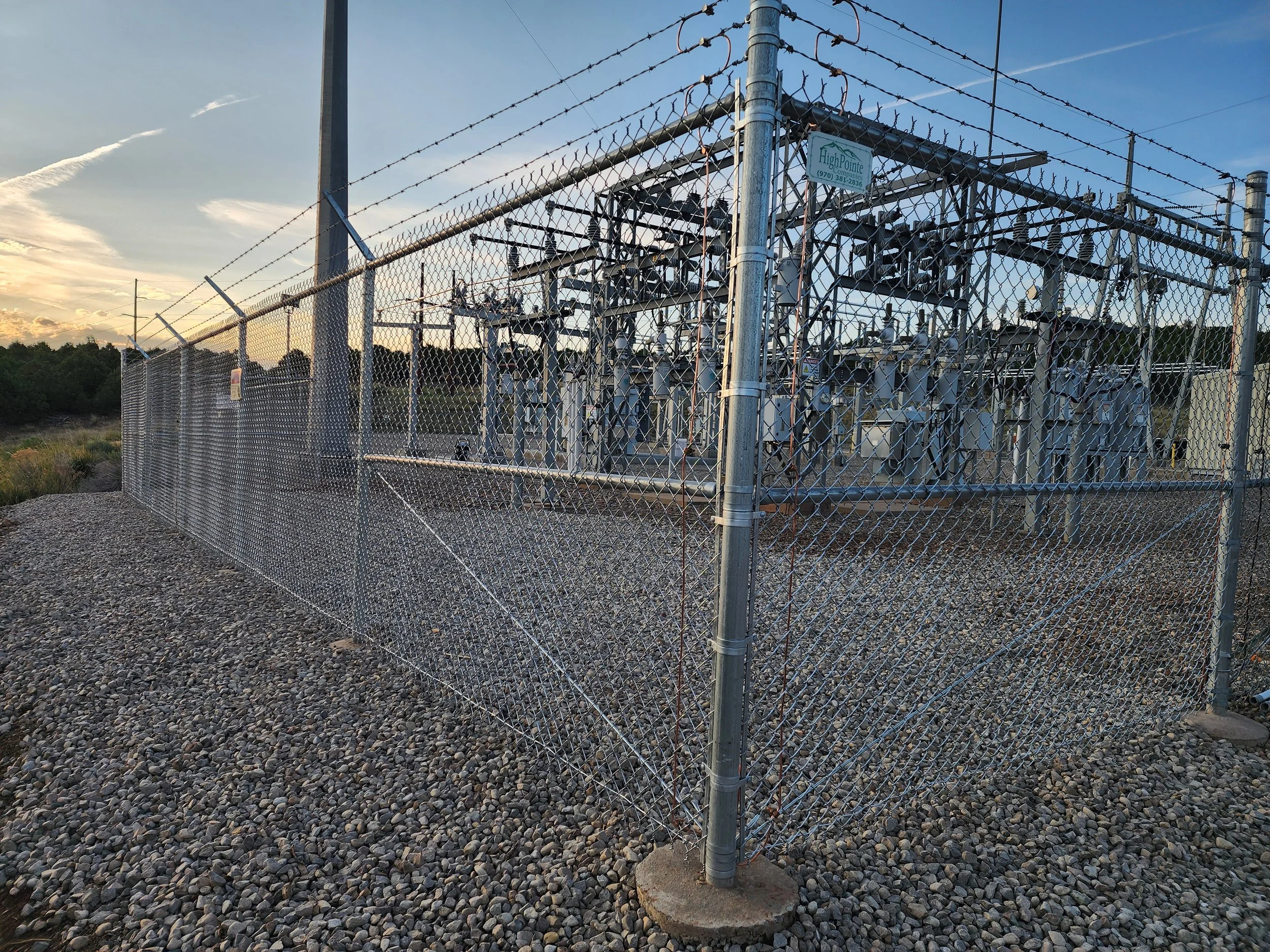 An electrical substation enclosed by a chain-link fence with barbed wire on top, situated on gravel ground under a sunset sky.