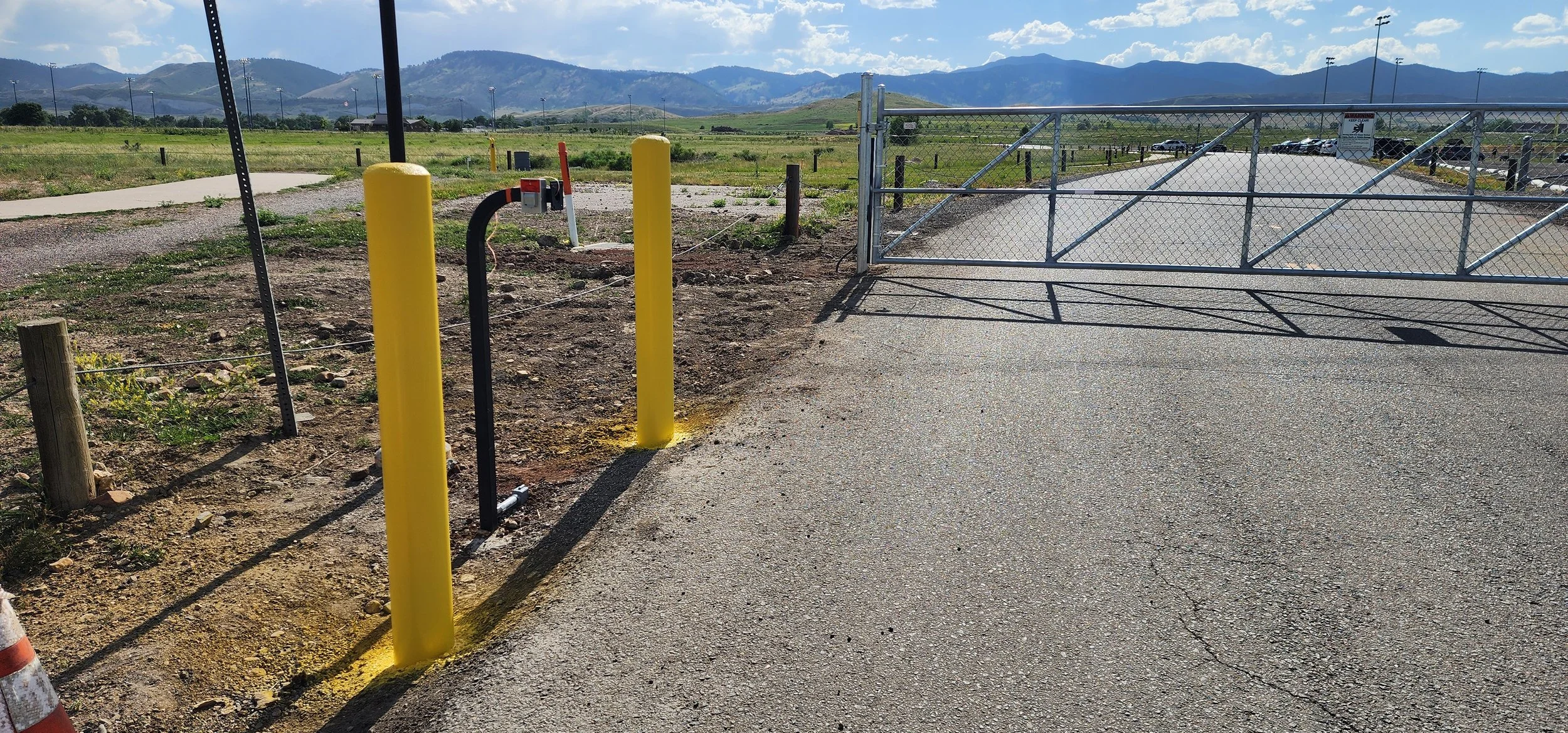 A gated parking lot with a chain-link gate, yellow safety posts, and a gravel path leading to the open gate, with mountains in the background.