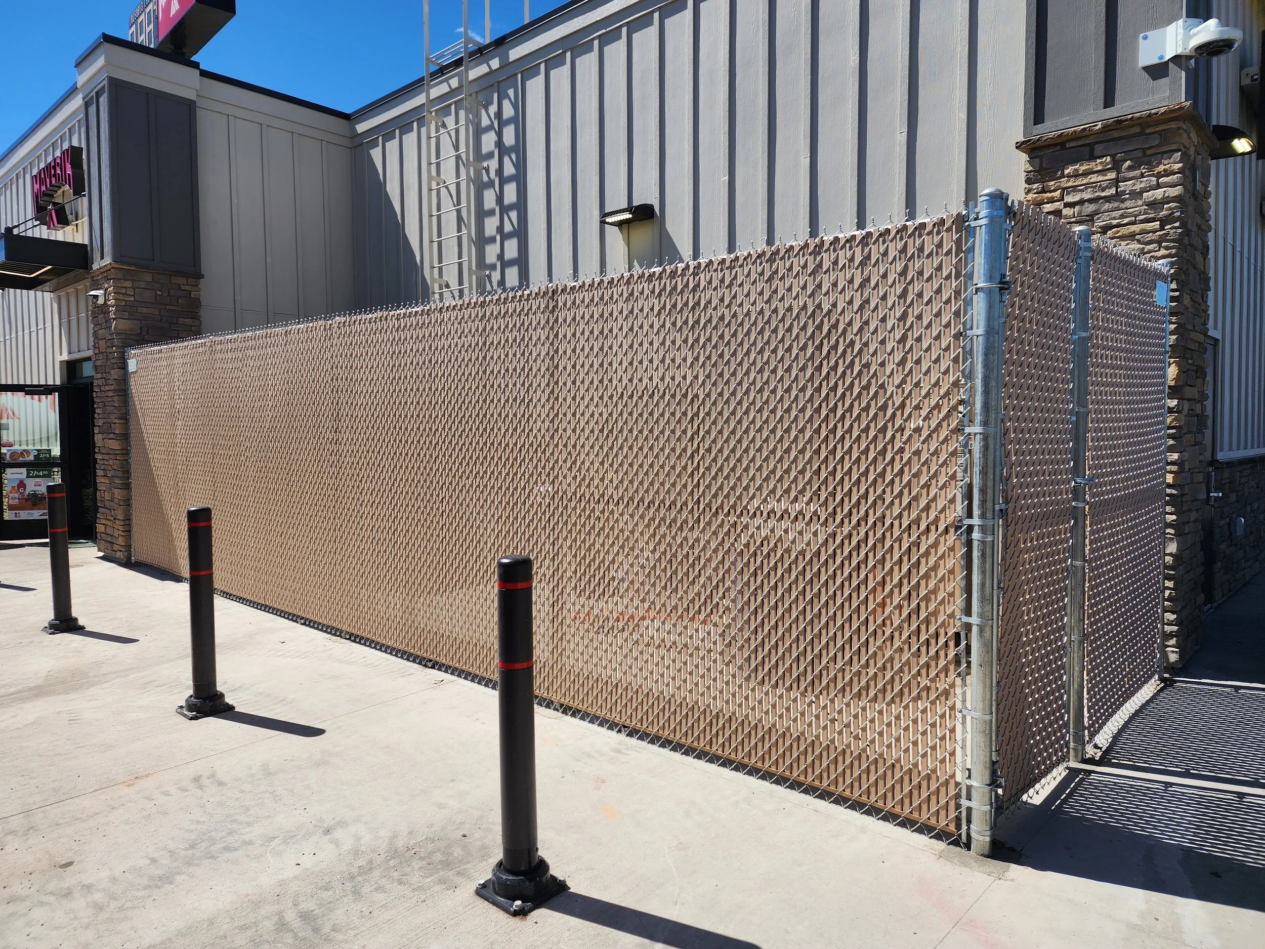 Chain-link fence with cardboard privacy screening on a sidewalk outside commercial buildings, with three black bollards in front and a corner of the building visible.