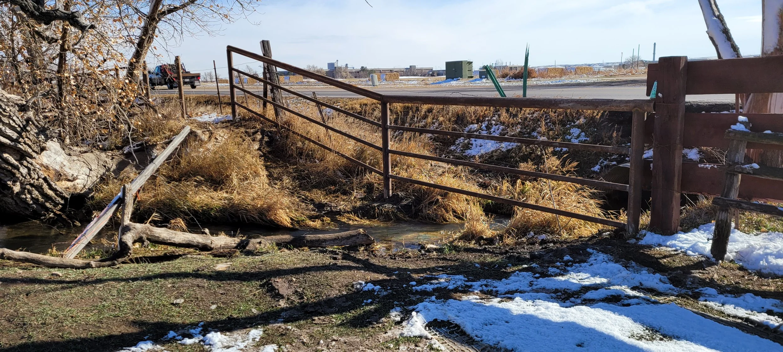 Fallen tree and damaged wooden fence near a small creek, with snow on the ground and an open field in the background.
