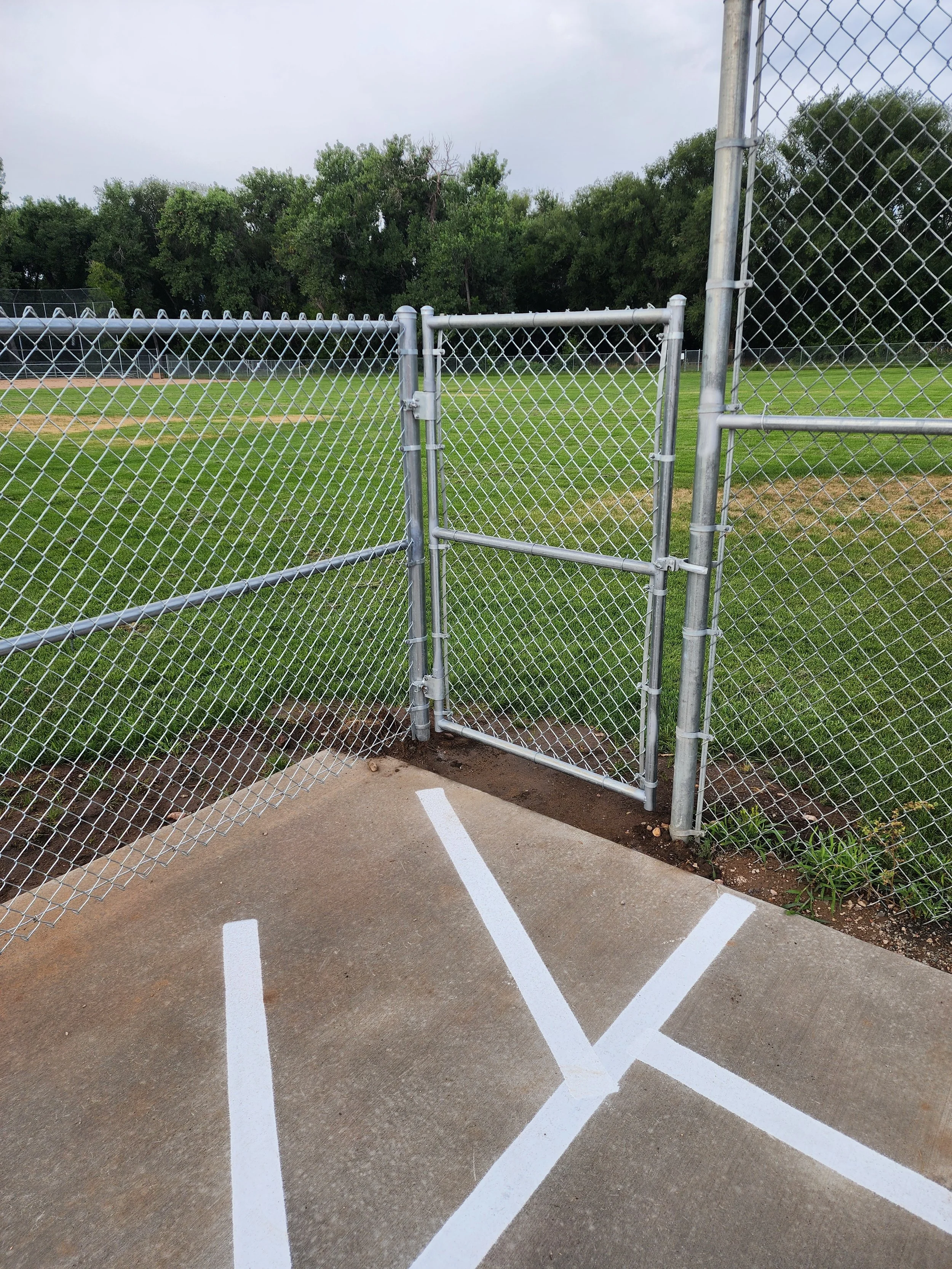 Closed chain-link fence gate at the entrance of a baseball or softball field, with painted parking lines on the concrete ground in the foreground, green grass, and trees in the background.