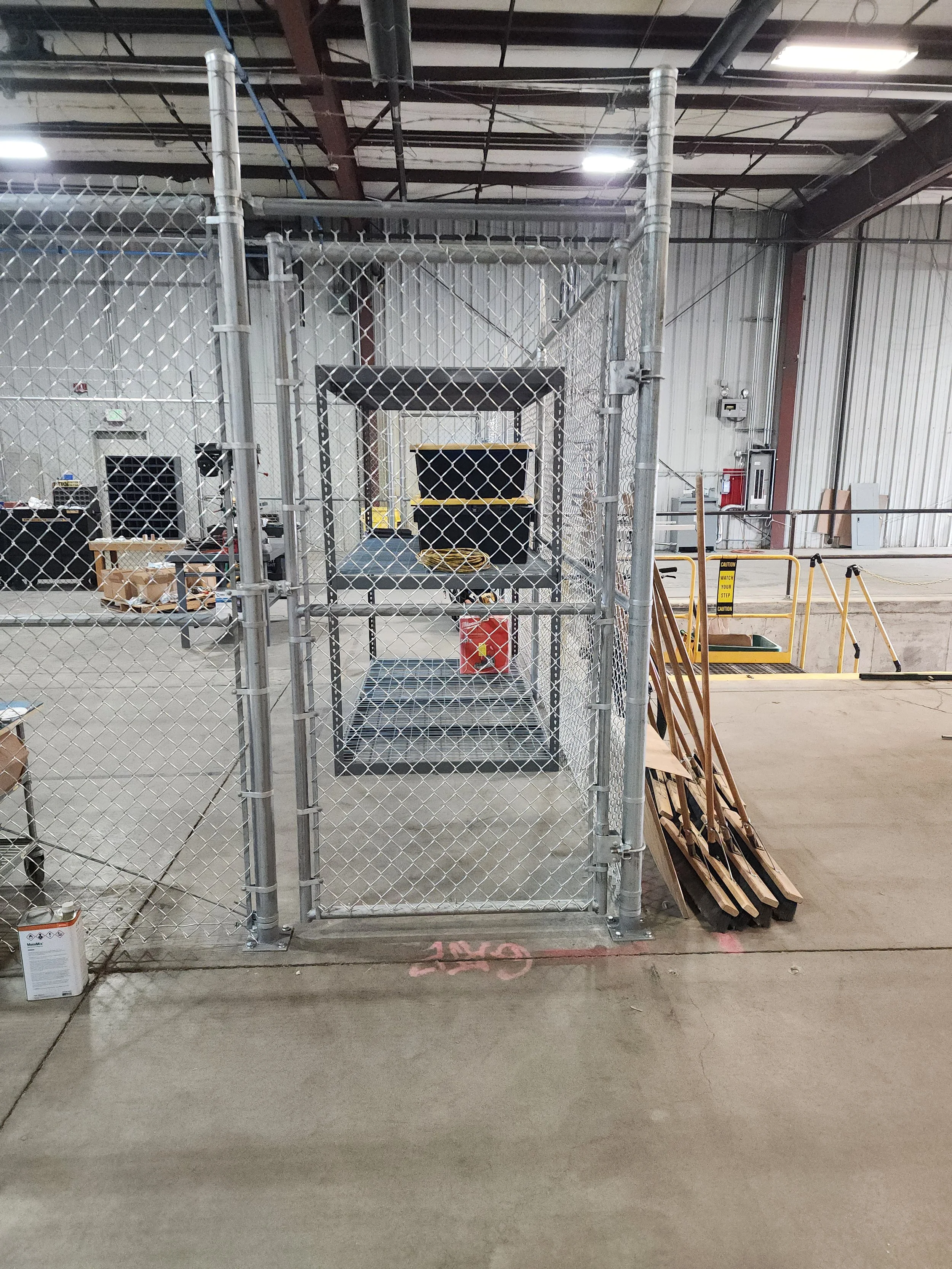 A fenced area inside an industrial or warehouse building with metal flooring and walls, containing construction or maintenance equipment including ladders, caution signs, and boxes. Fluorescent lights illuminate the area.