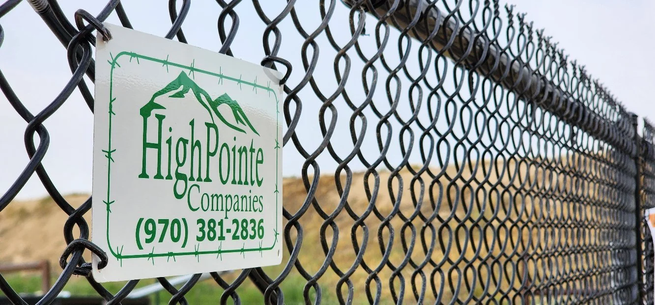 A close-up view of a chain-link fence with a High Pointe Companies sign attached, displaying a phone number, against a background of rolling hills under a cloudy sky.